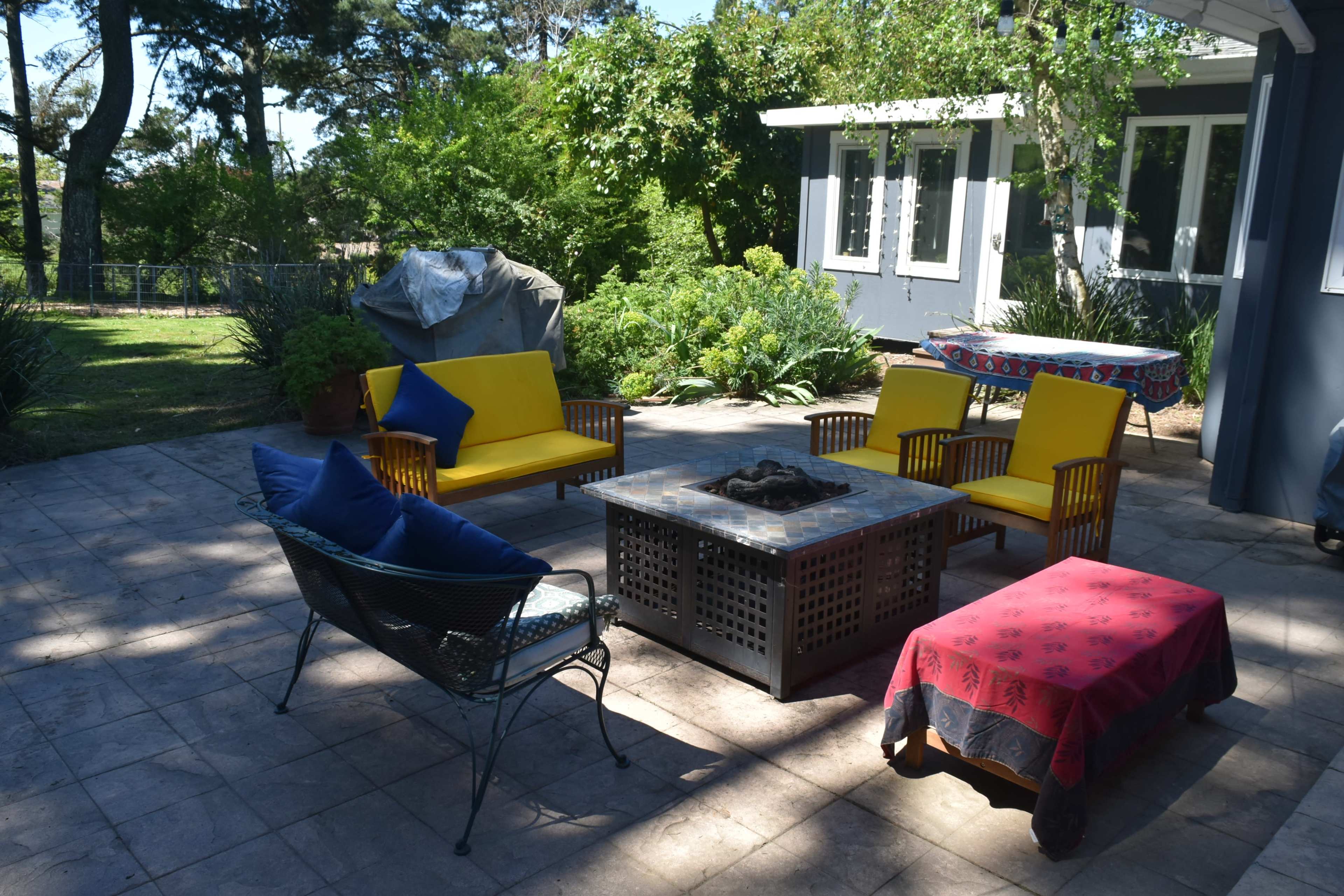 A patio features a rectangular fire pit surrounded by yellow and blue seating, with greenery and a house in the background.