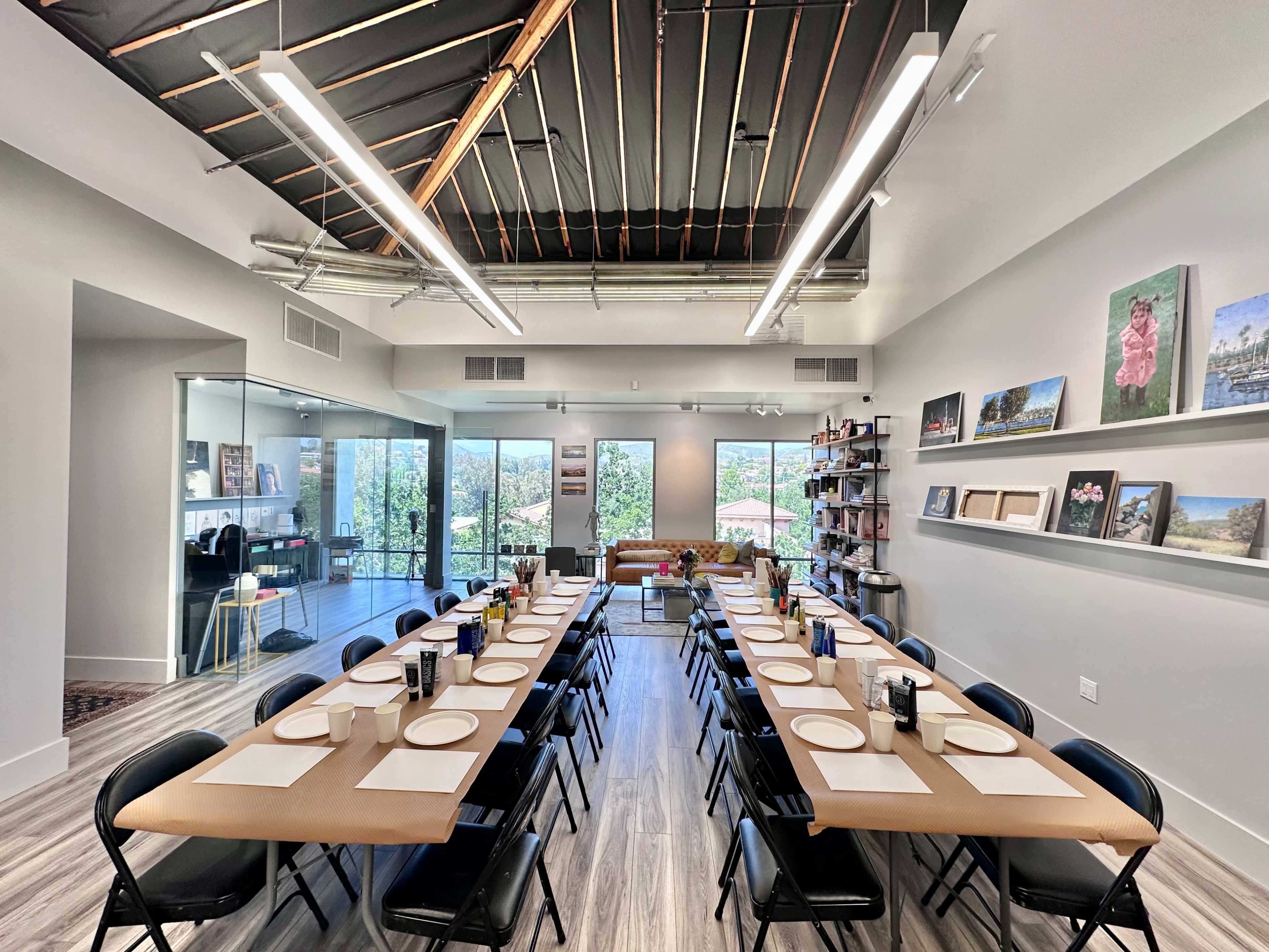 A modern dining room features a long table set with plates and utensils, flanked by black chairs, and large windows showcasing a view outside.