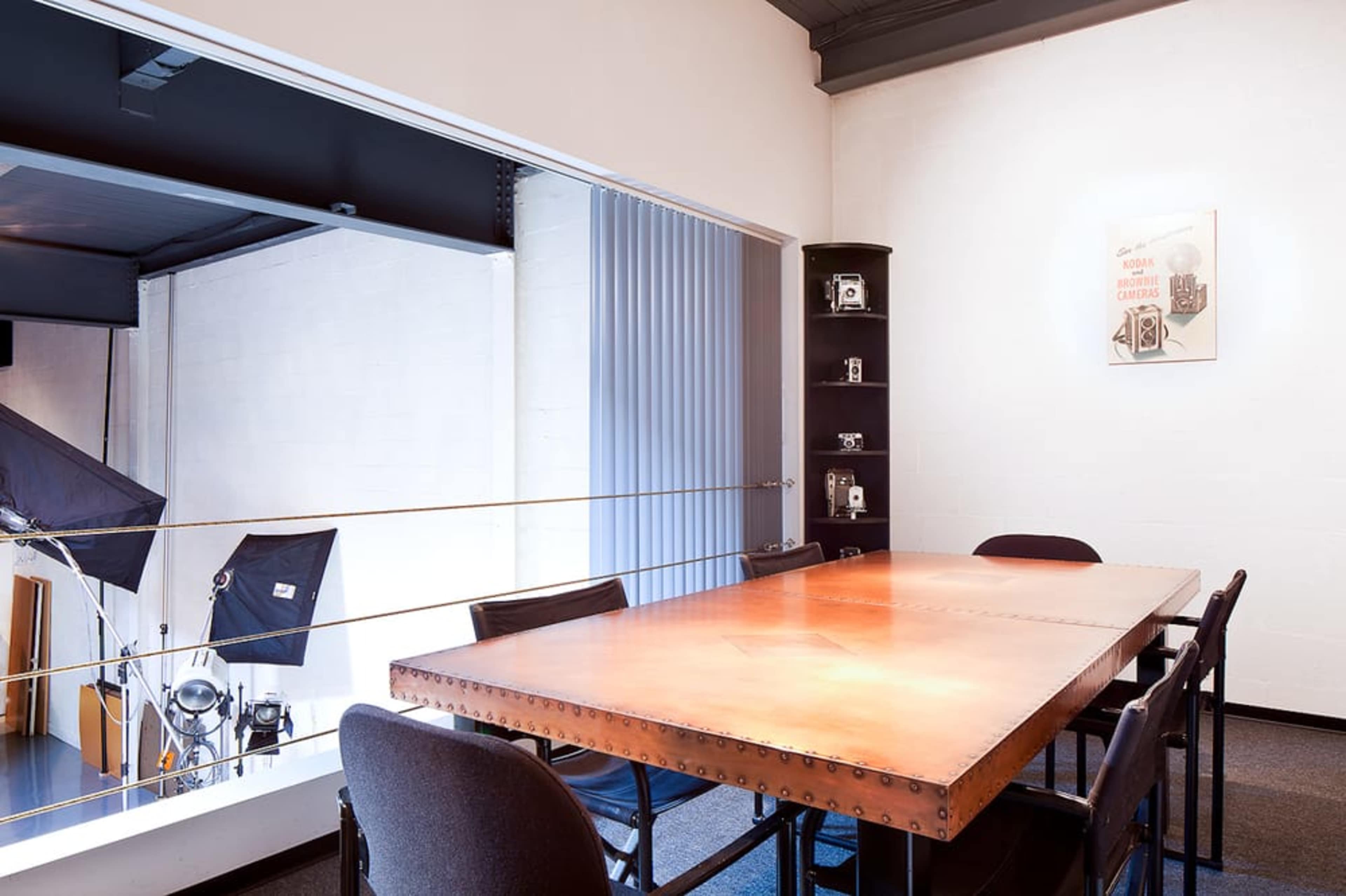 A large wooden table is surrounded by black chairs in a studio-like meeting room with a balcony area and photography equipment visible in the background.