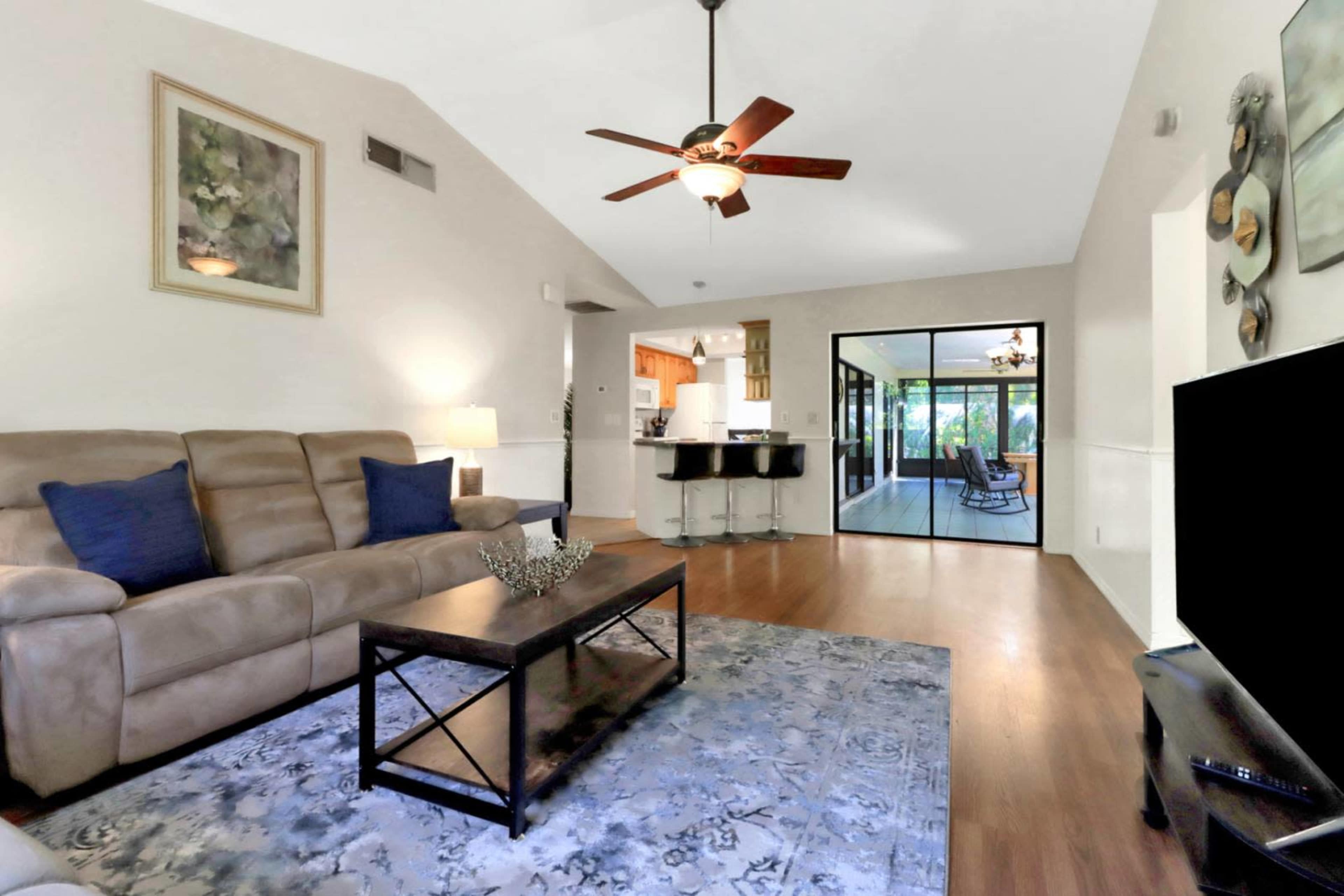 The image shows a living room with a brown leather sofa, a coffee table, and a ceiling fan, leading to a glass-walled area that appears to be a sunroom or porch.