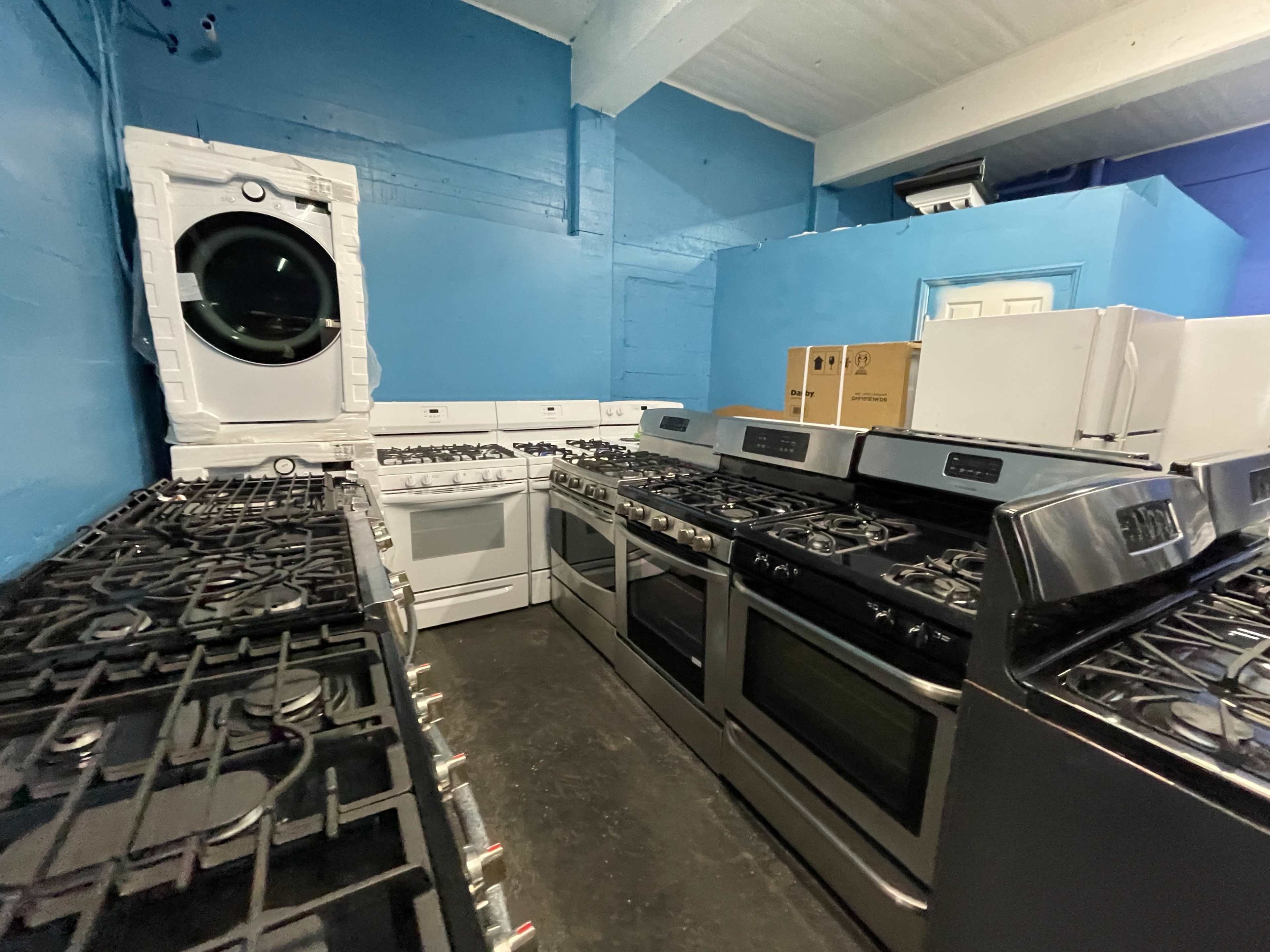 The image shows a room featuring multiple kitchen appliances, including several gas ranges and a washer and dryer against blue-painted walls.