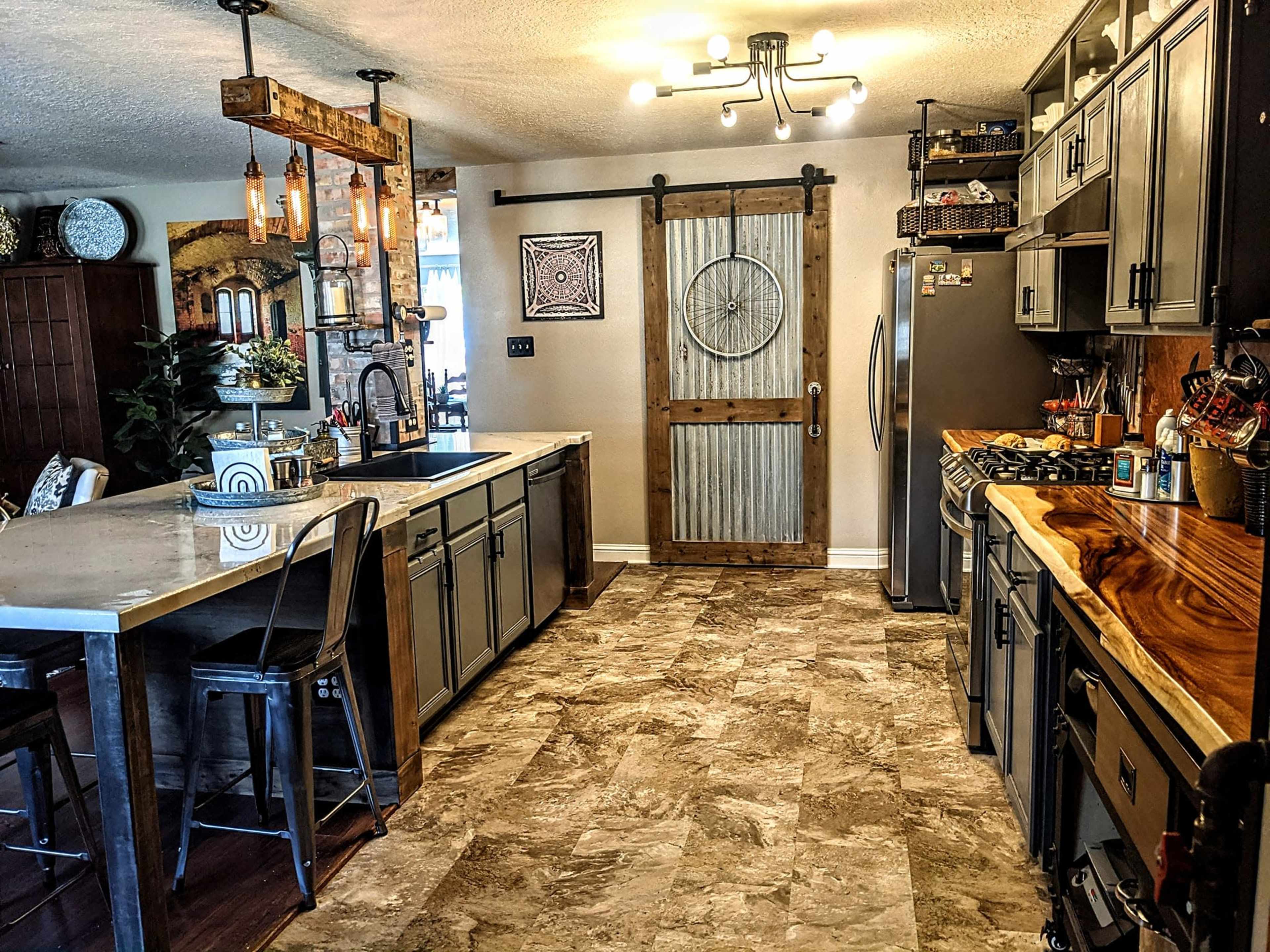 The image depicts a modern kitchen with a large island, stainless steel appliances, a sliding barn door, and a mix of open and closed cabinetry.