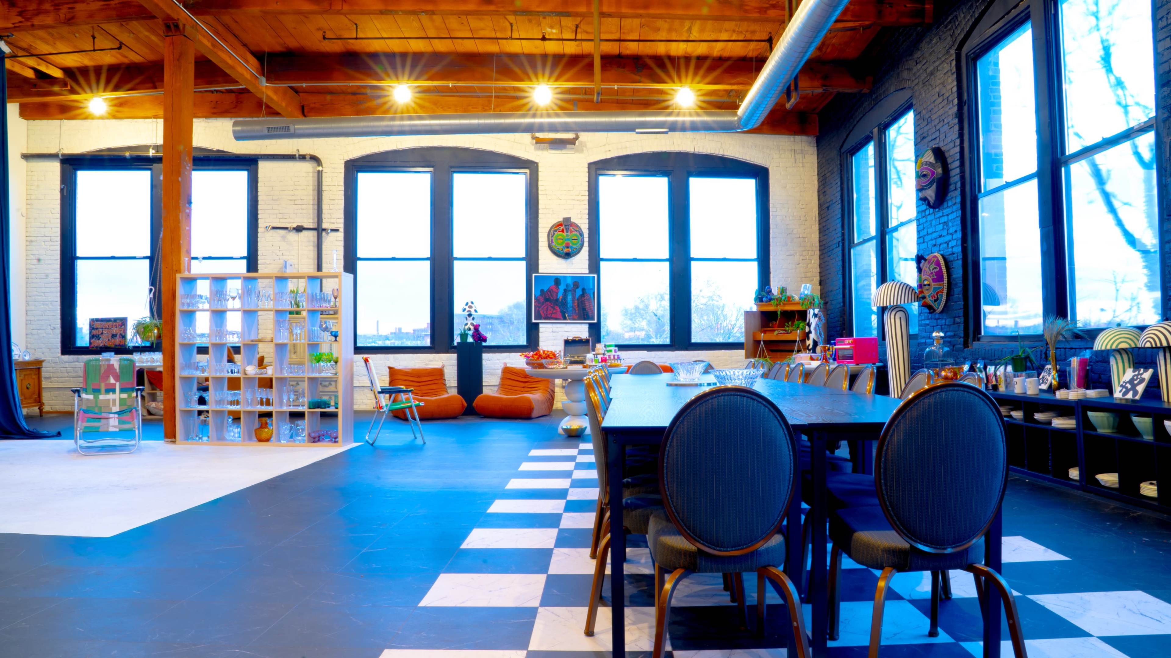 The image depicts a spacious room with large windows, wooden beams, a long dining table, colorful seating, and various decorative elements.