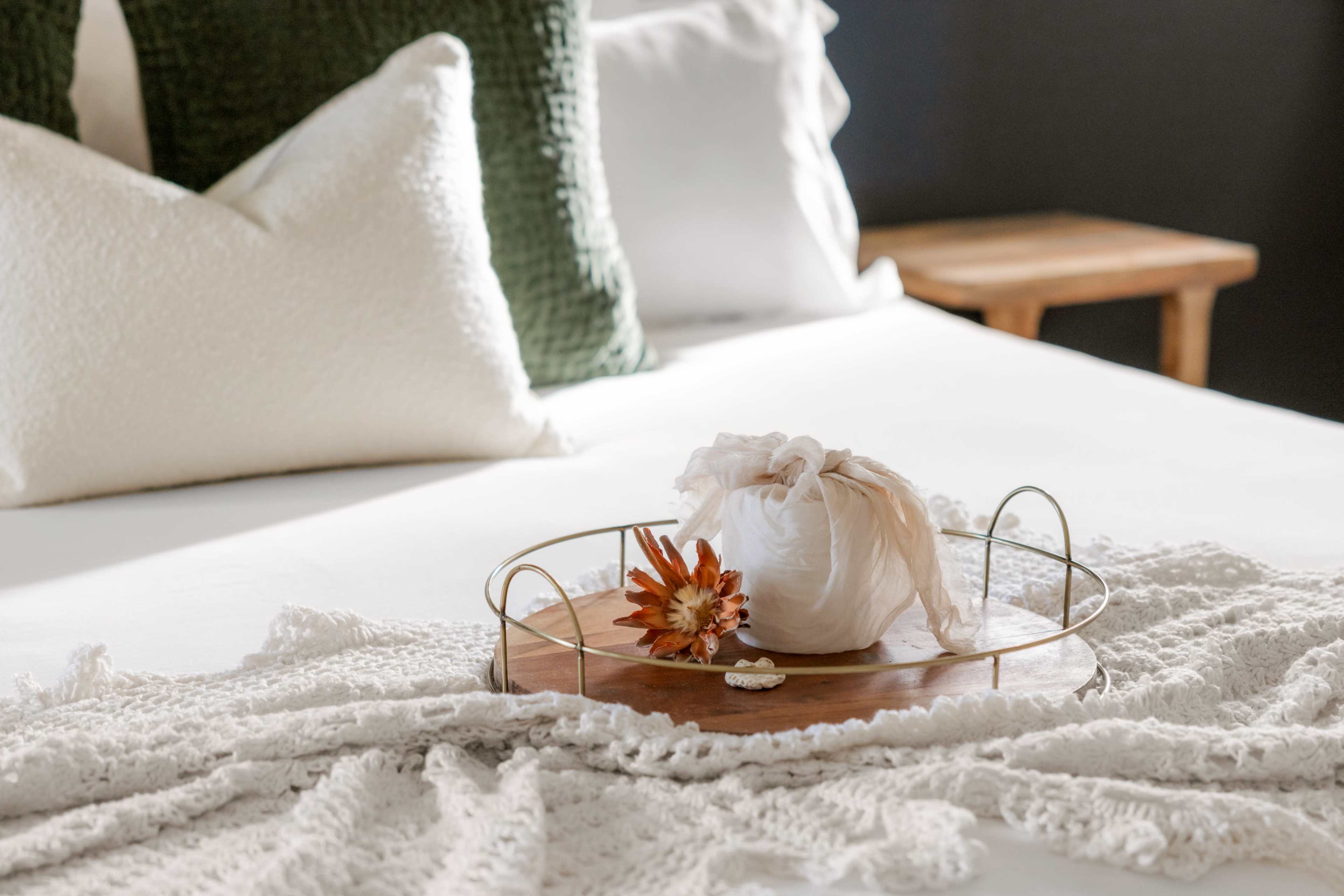 A wooden tray with a wrapped item and a flower sits on a neatly made bed adorned with pillows and a textured blanket.
