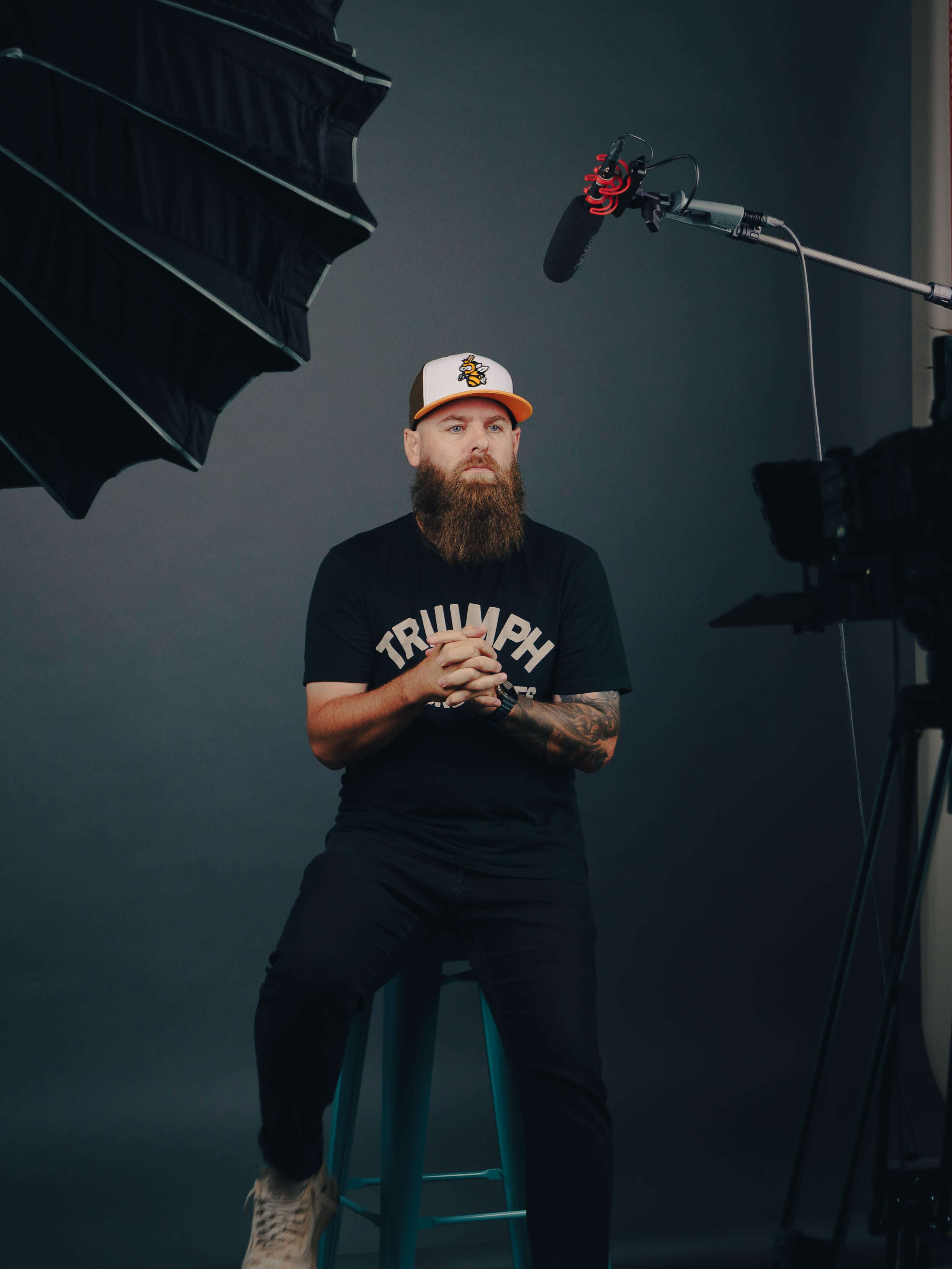 A man with a beard sits on a stool in front of a camera setup, wearing a black shirt and a baseball cap, with large studio lights overhead.
