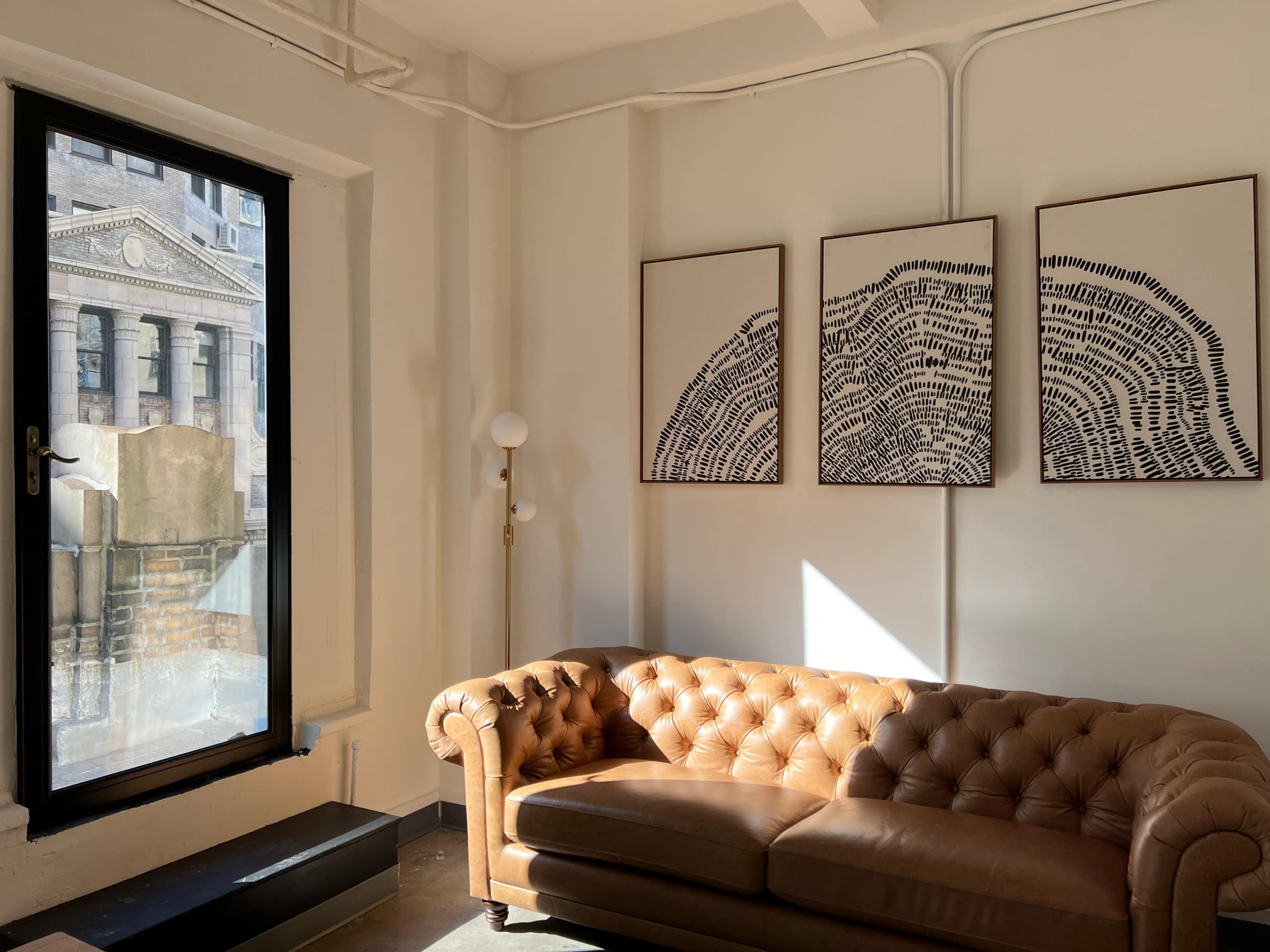 The image shows a sunlit room featuring a brown leather chesterfield sofa and three framed abstract artworks on the wall, with a large window revealing a view of a building exterior.