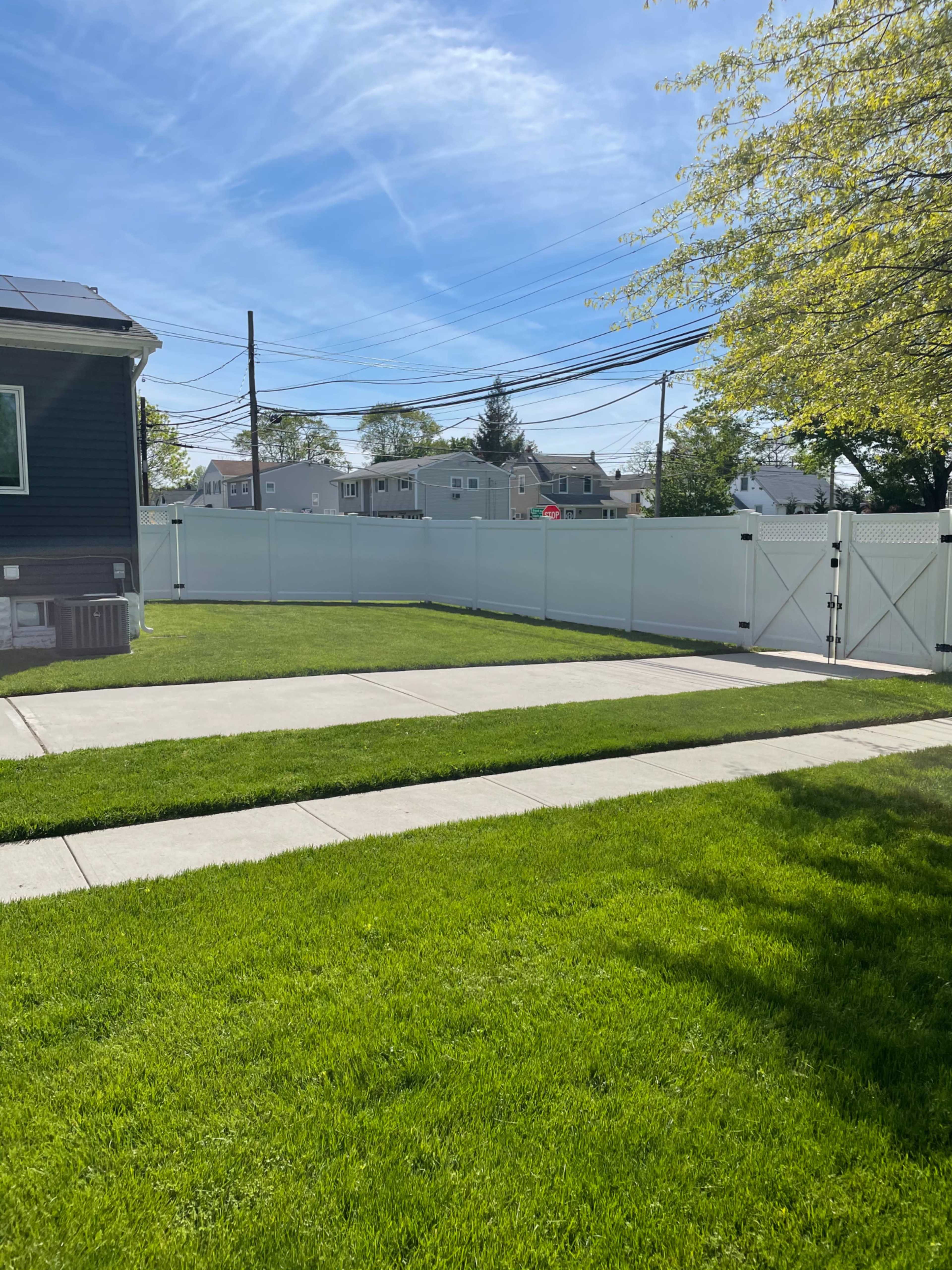 The image shows a well-maintained backyard with green grass, concrete pathways, and a white vinyl fence in the background.