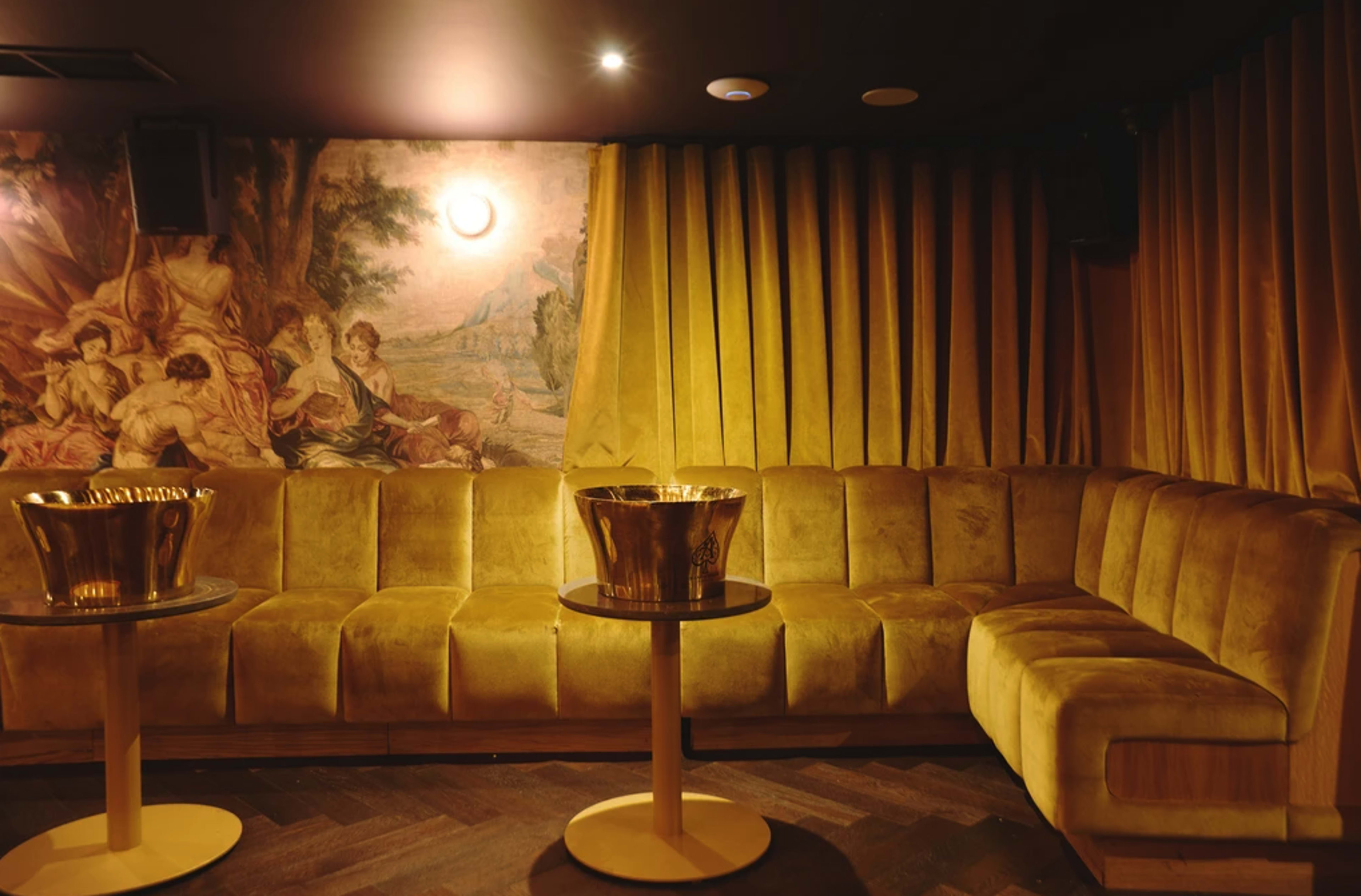 The image depicts a dimly lit lounge with a plush, golden-yellow velvet sofa, circular tables, and a painted mural on the wall.