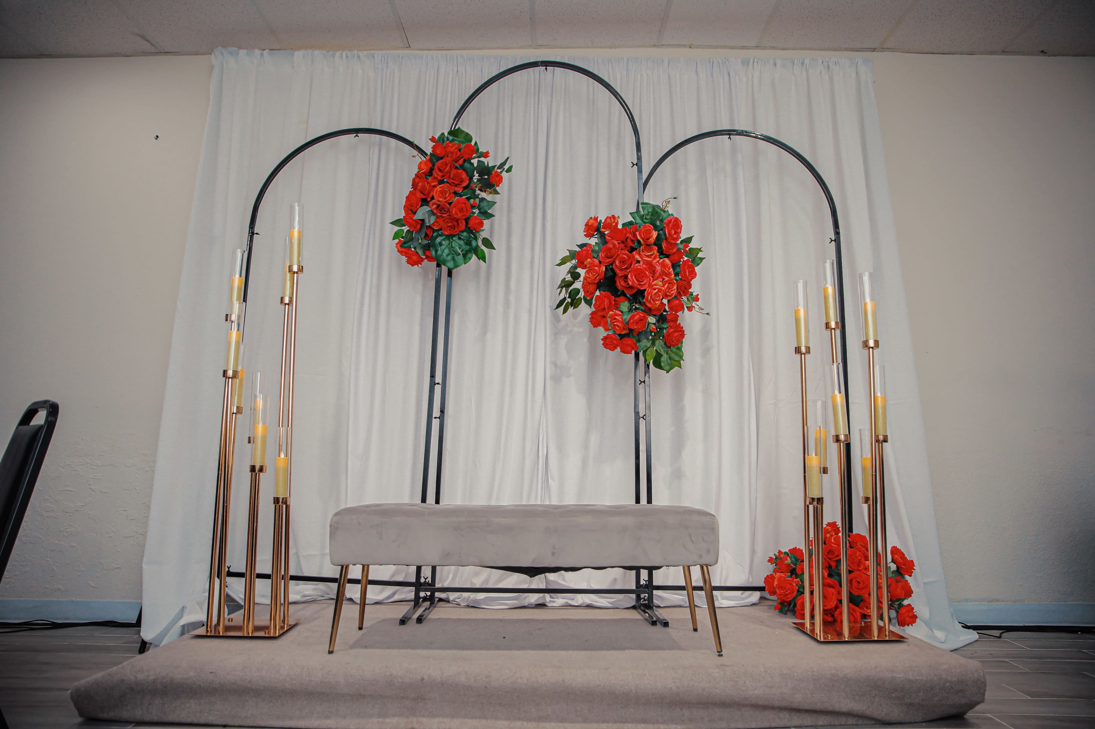 A floral archway with red flowers is set up against a white backdrop, flanked by tall gold candleholders.