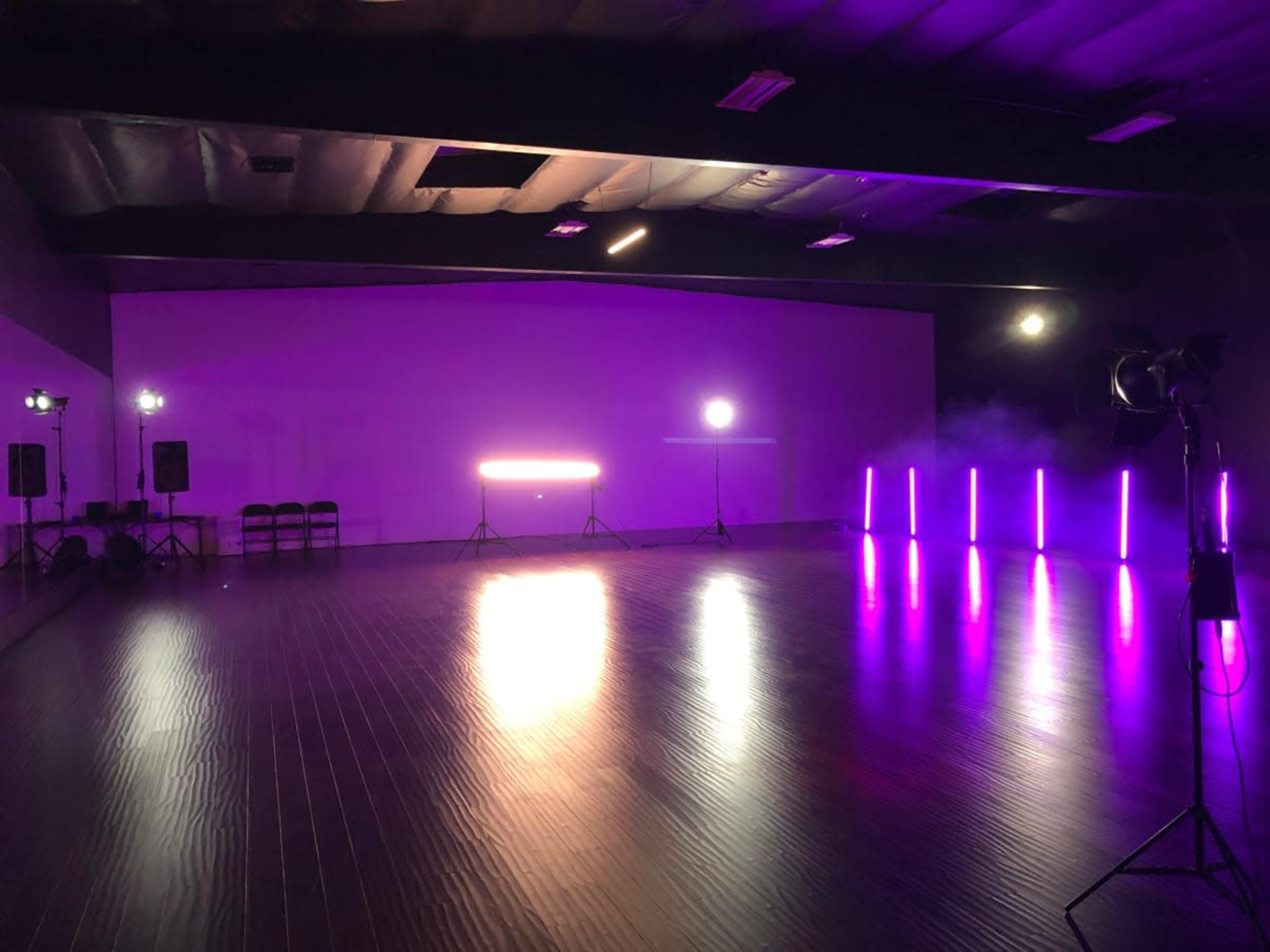 A spacious studio is illuminated by purple lighting and equipped with audio speakers and multiple light stands.