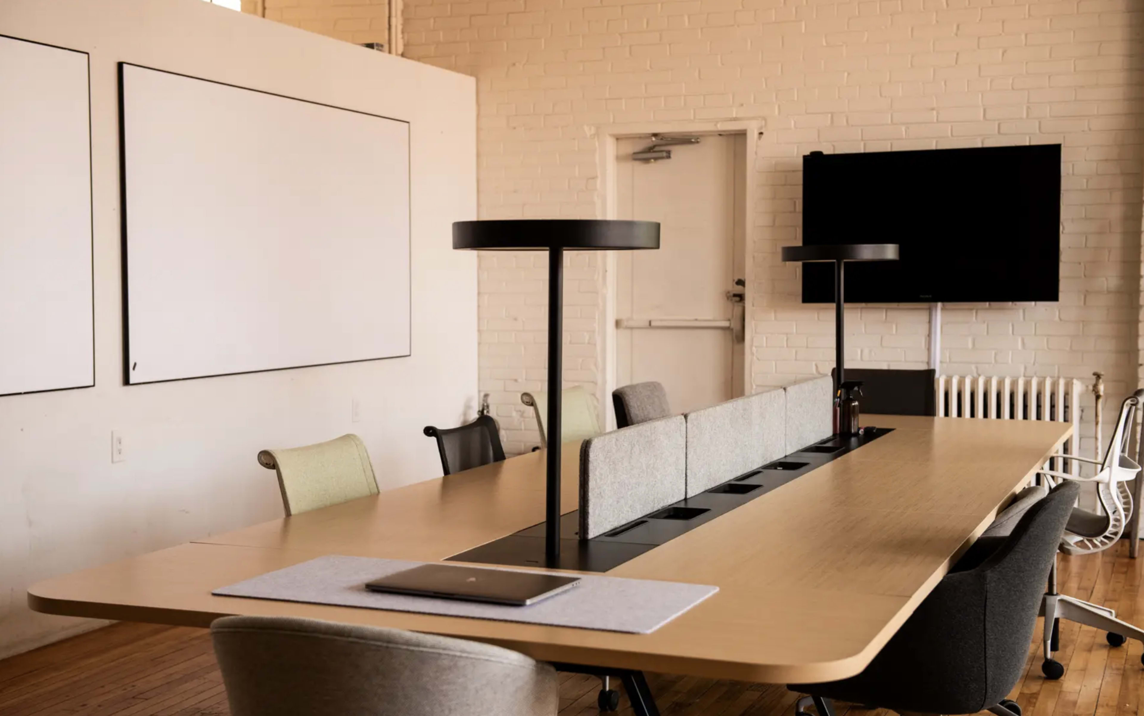 A modern conference room features a long wooden table, stylish chairs, and a large screen mounted on the wall.
