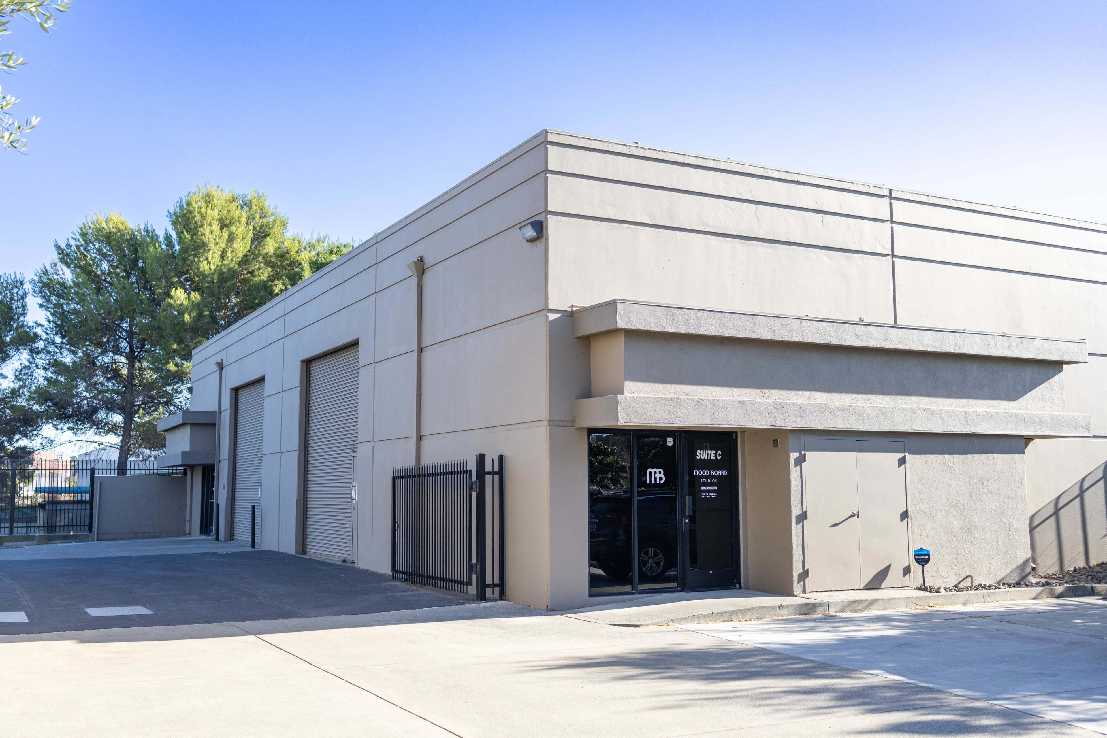 The image shows a light-colored commercial building with a modern facade, featuring large roll-up doors and an entrance with signage.