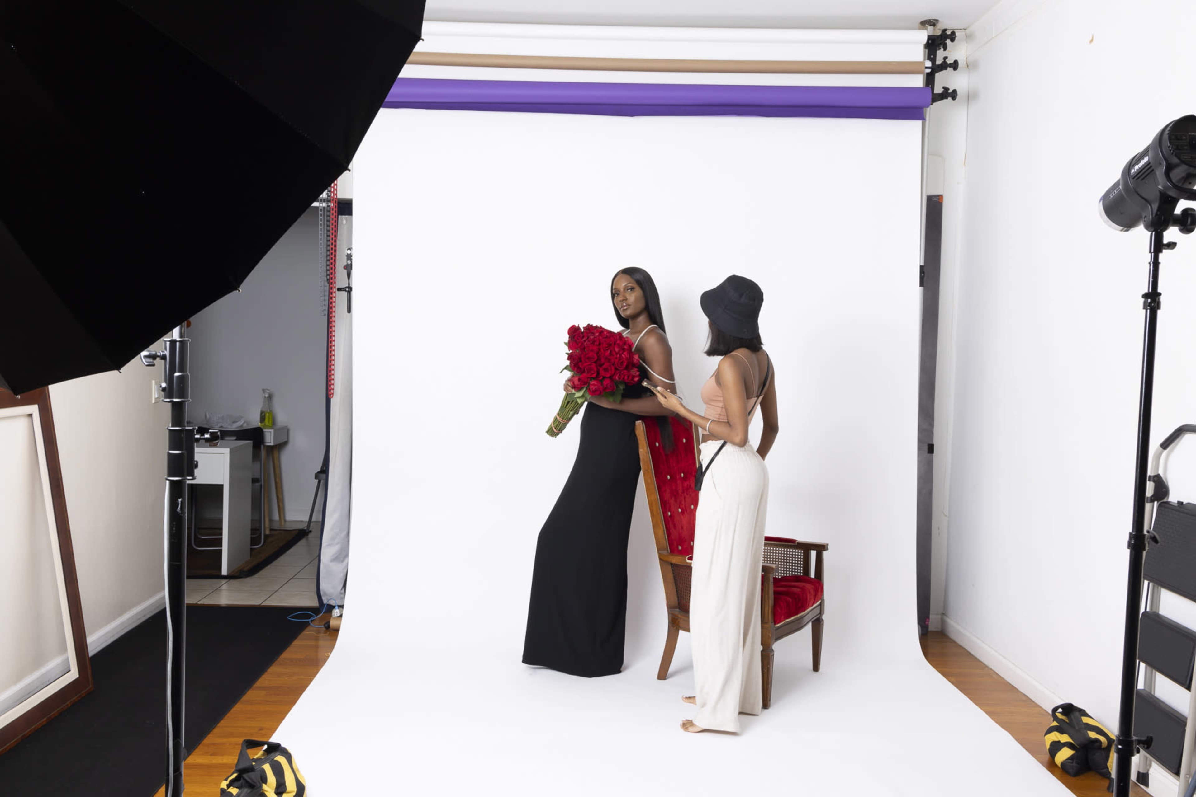 A model in a black dress poses with a large bouquet of red roses while another person adjusts her outfit in a photo studio setting.