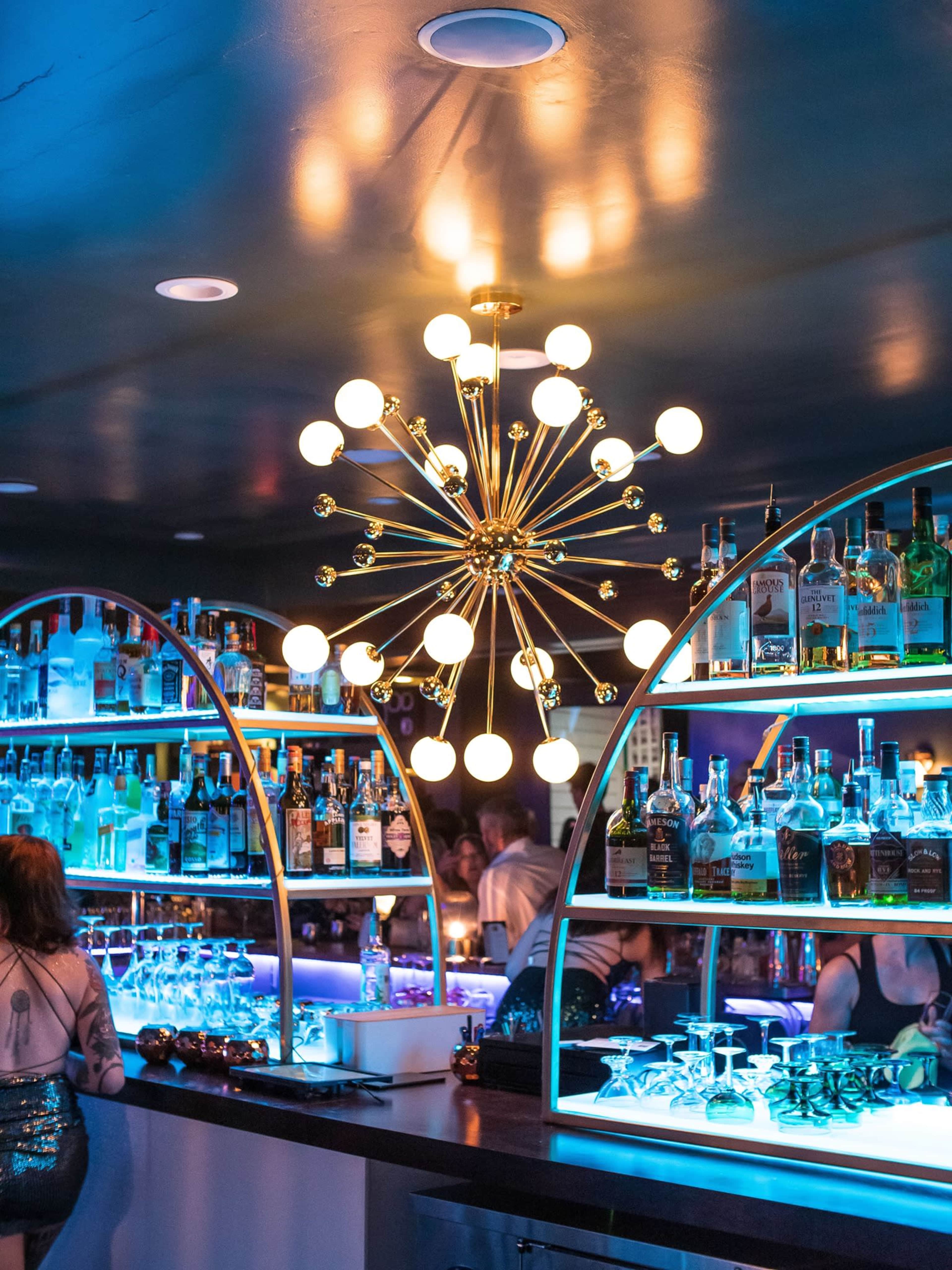 A stylish bar with illuminated shelves displaying various bottles of liquor and a large, modern chandelier above.