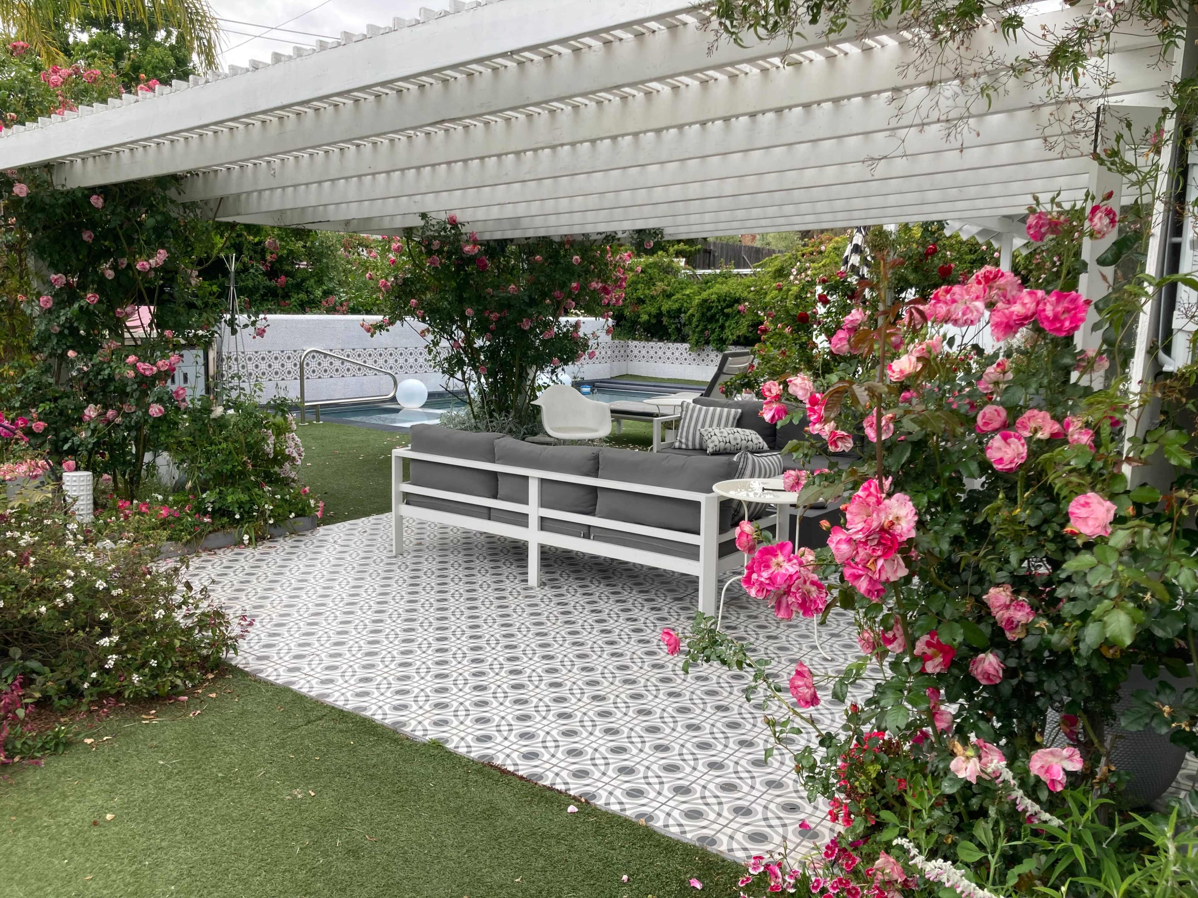 A covered seating area surrounded by flowering plants and a patterned tile floor leads to a pool in a landscaped backyard.