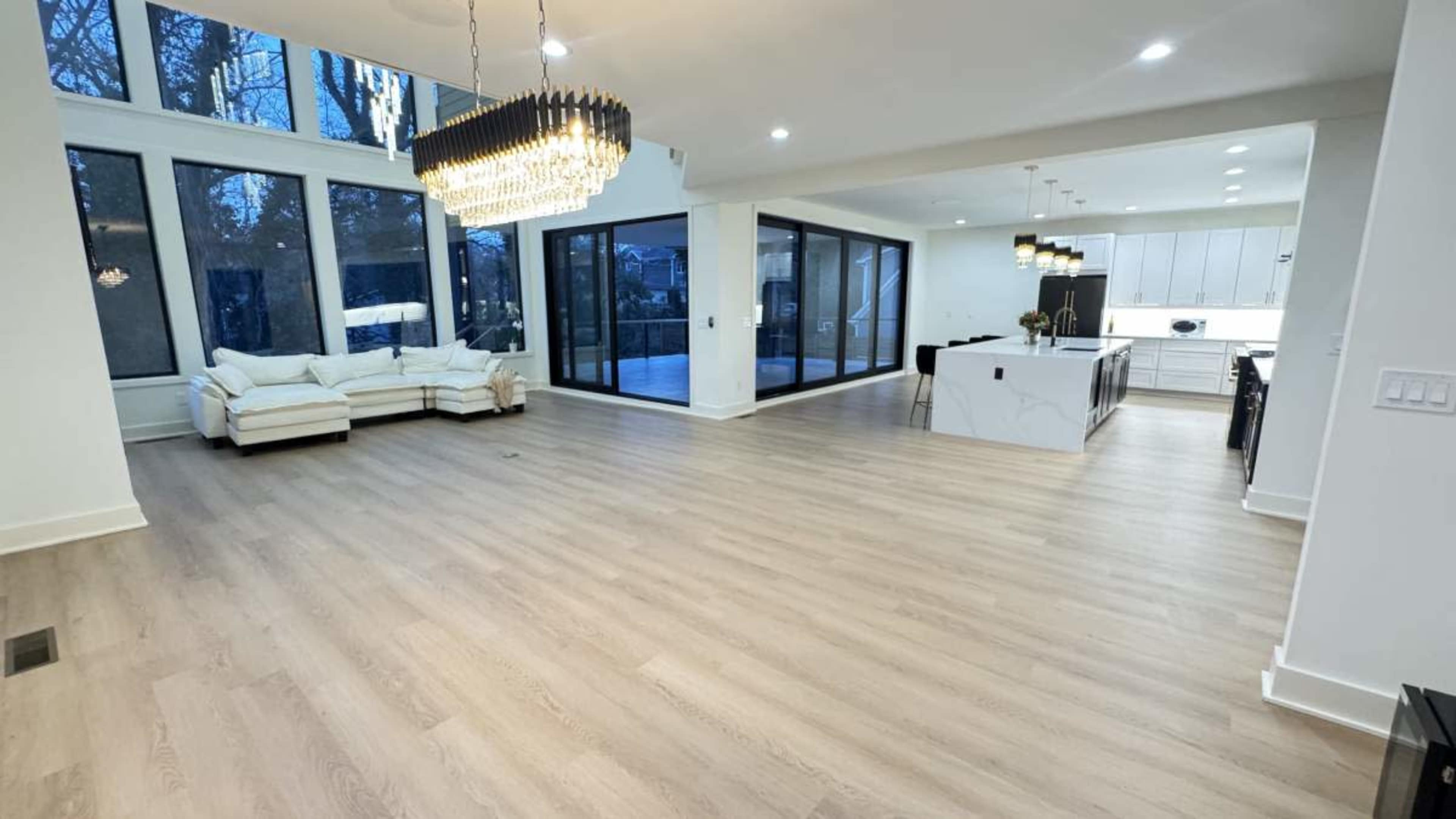 The image shows a spacious, modern living area with large windows, a white sofa, and an open kitchen layout.