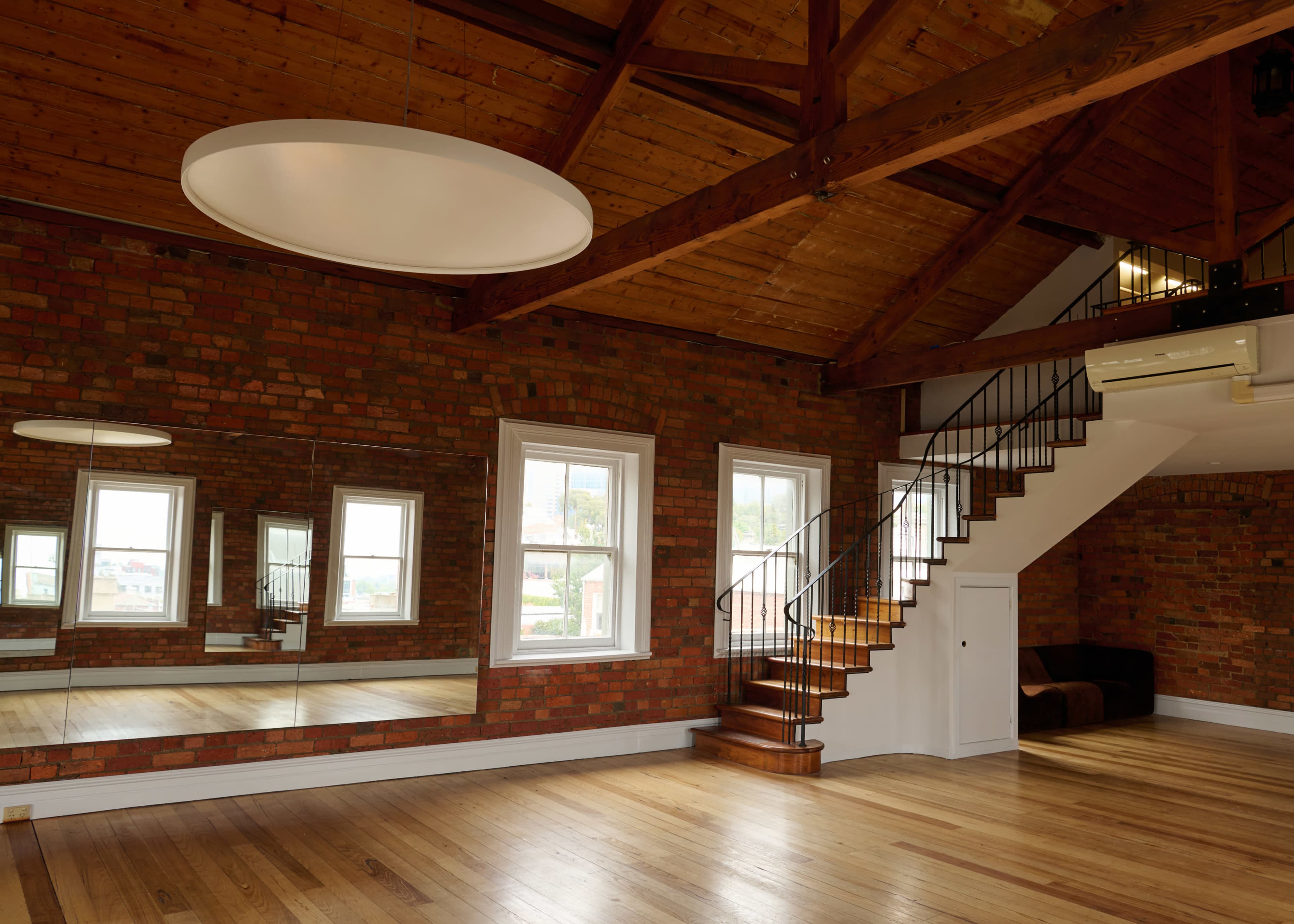 The spacious interior features exposed brick walls, large windows, and a staircase leading to a mezzanine level.