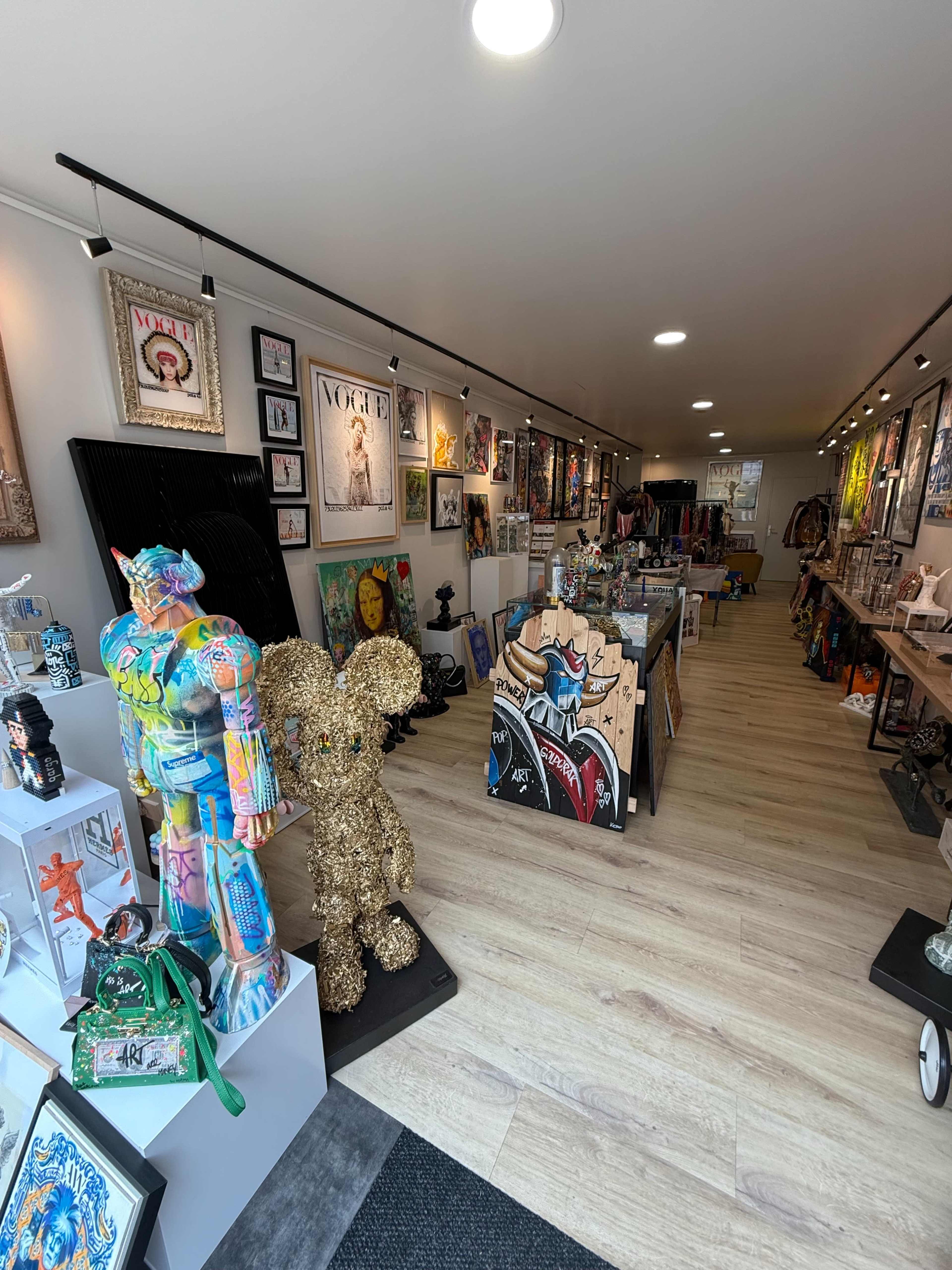 The image shows the interior of an art gallery featuring a variety of colorful artwork, sculptures, and decorative items displayed along the walls and on various shelves.