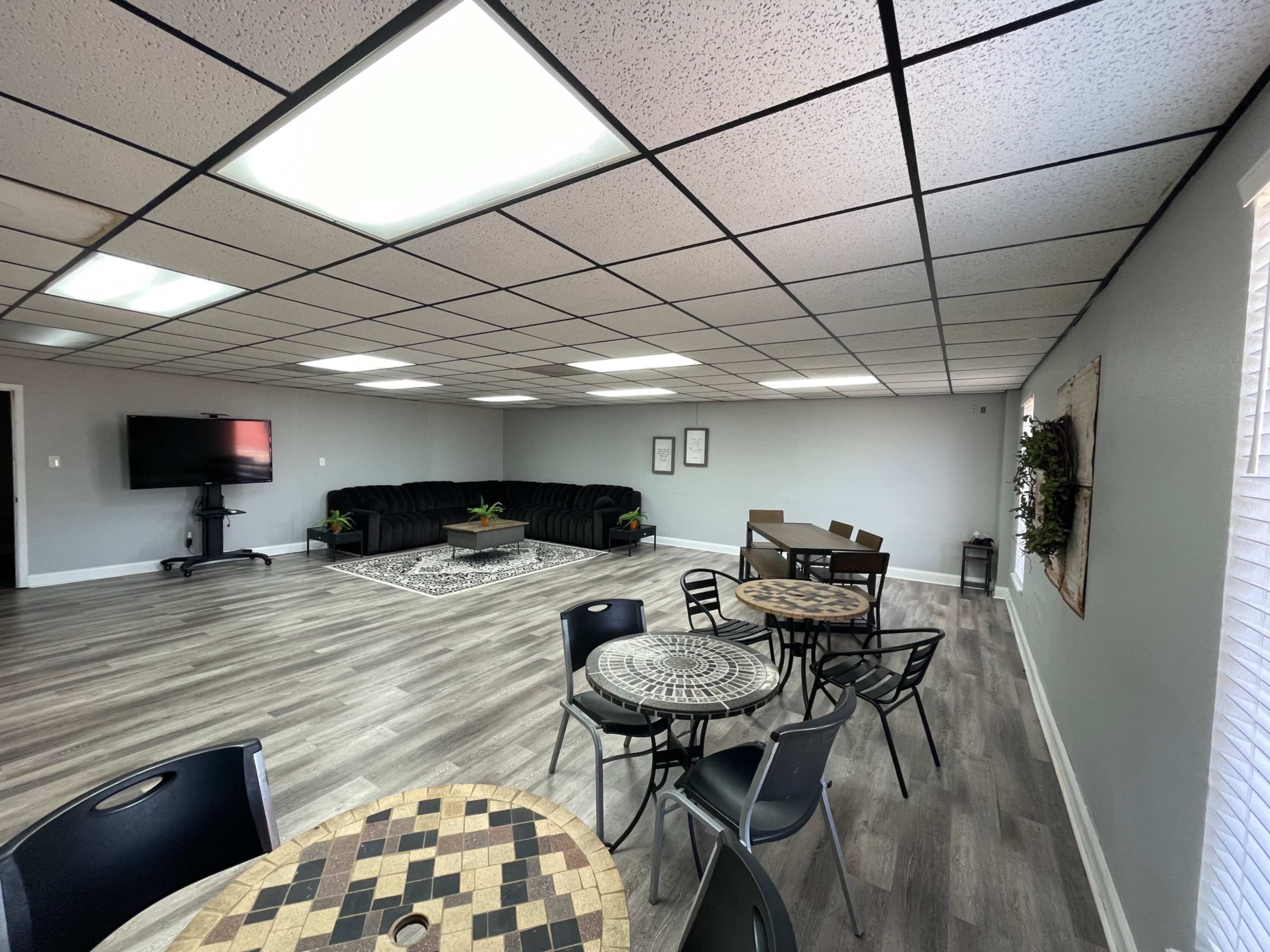 The image shows a spacious room with a seating area featuring black couches, a television mounted on the wall, and several round tables with chairs.