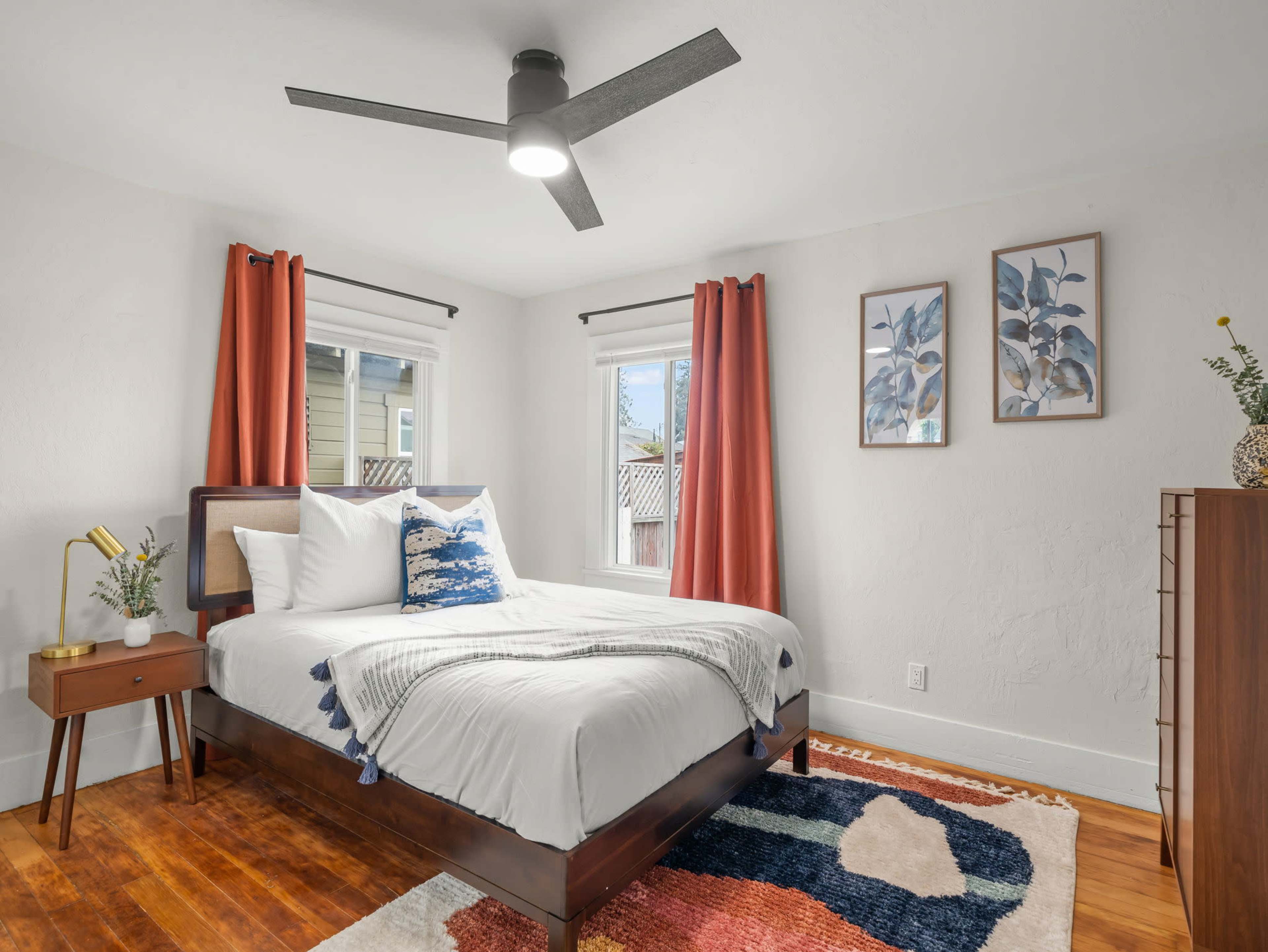 A well-lit bedroom features a bed with white linens, orange curtains on two windows, and decorative wall art.
