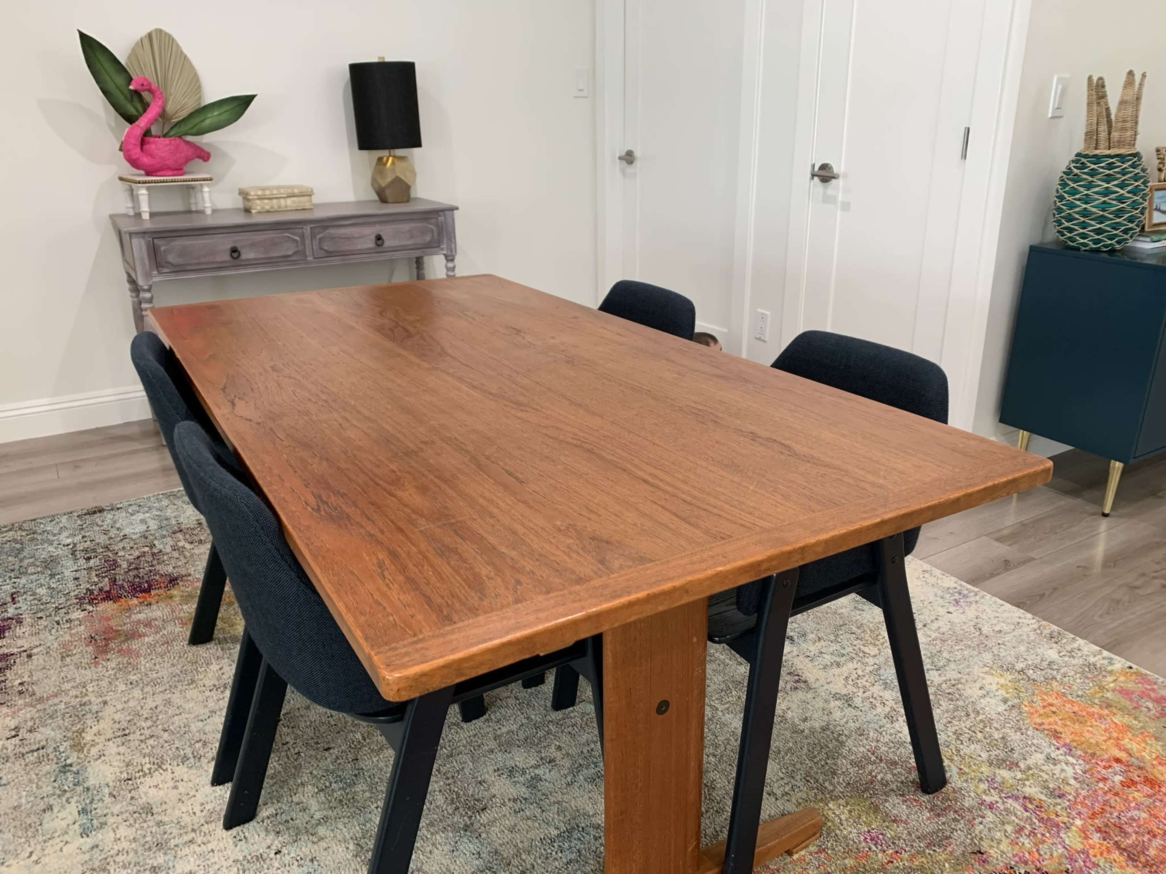 A rectangular wooden dining table is surrounded by four navy blue chairs in a brightly lit room with a decorative side table and colorful rug.