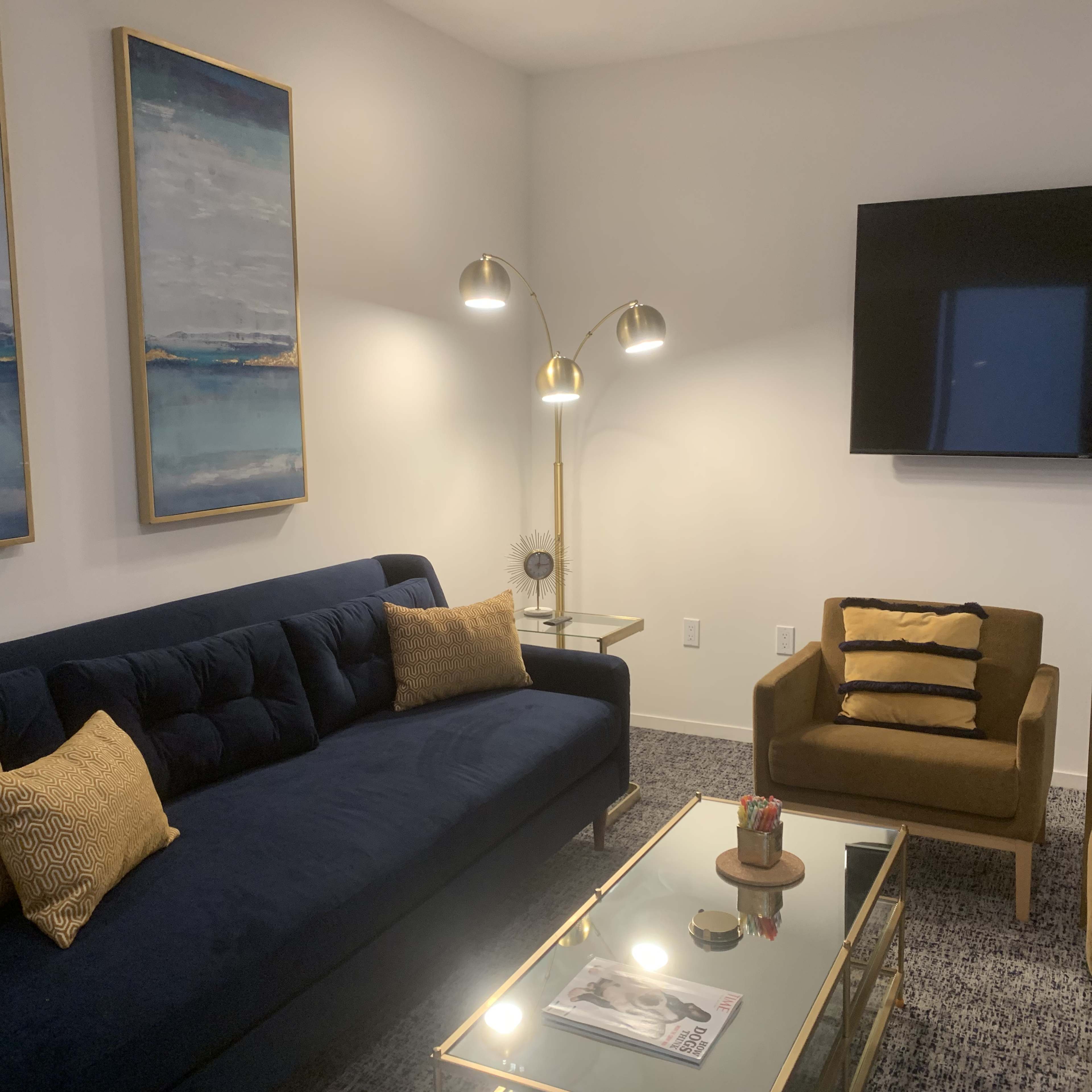 A modern living room featuring a blue sofa, a brown armchair, a gold lamp, and a glass coffee table with decorative items.
