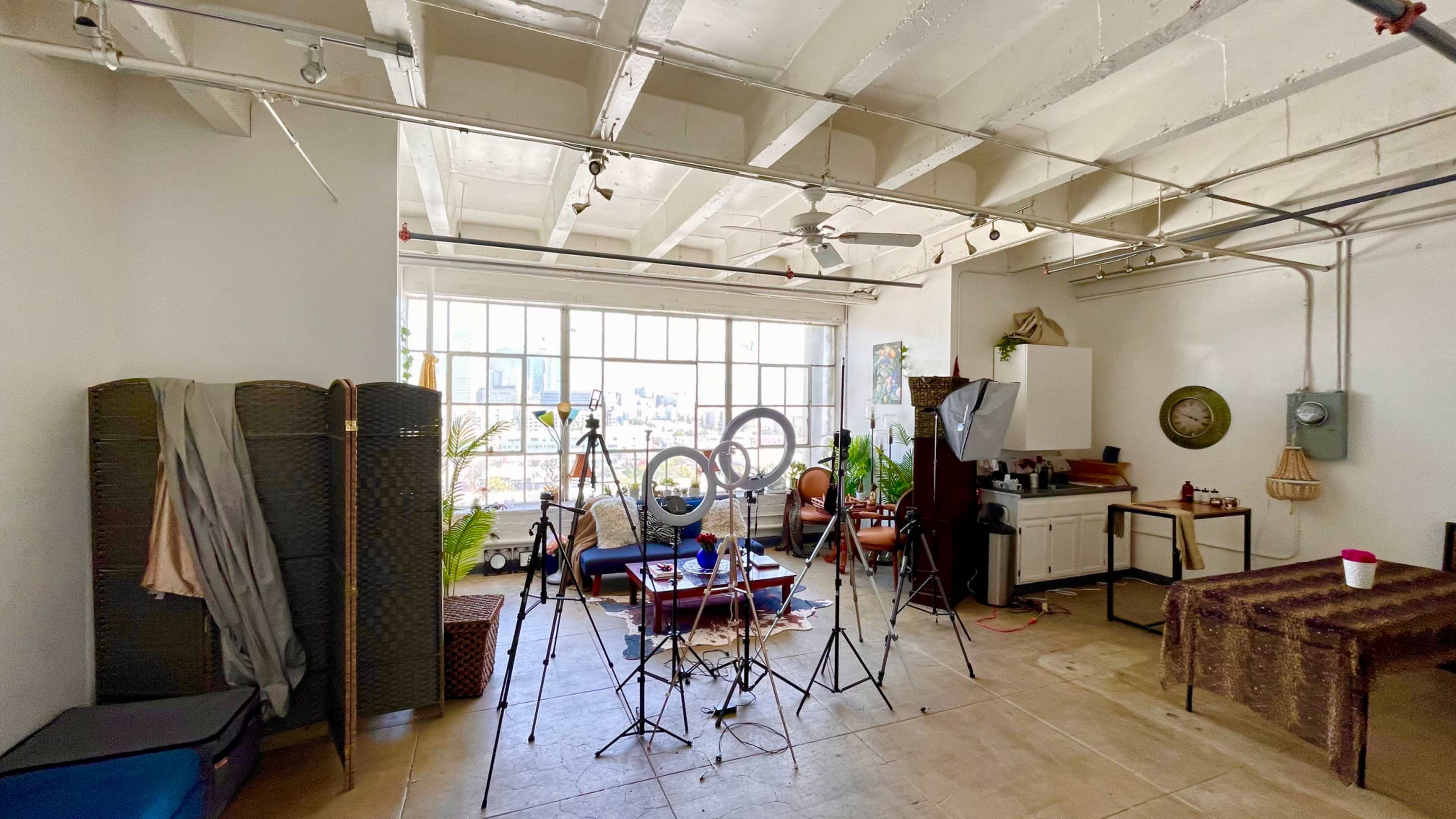 A spacious room with large windows features various photography equipment, green plants, and furniture arranged for a creative setup.