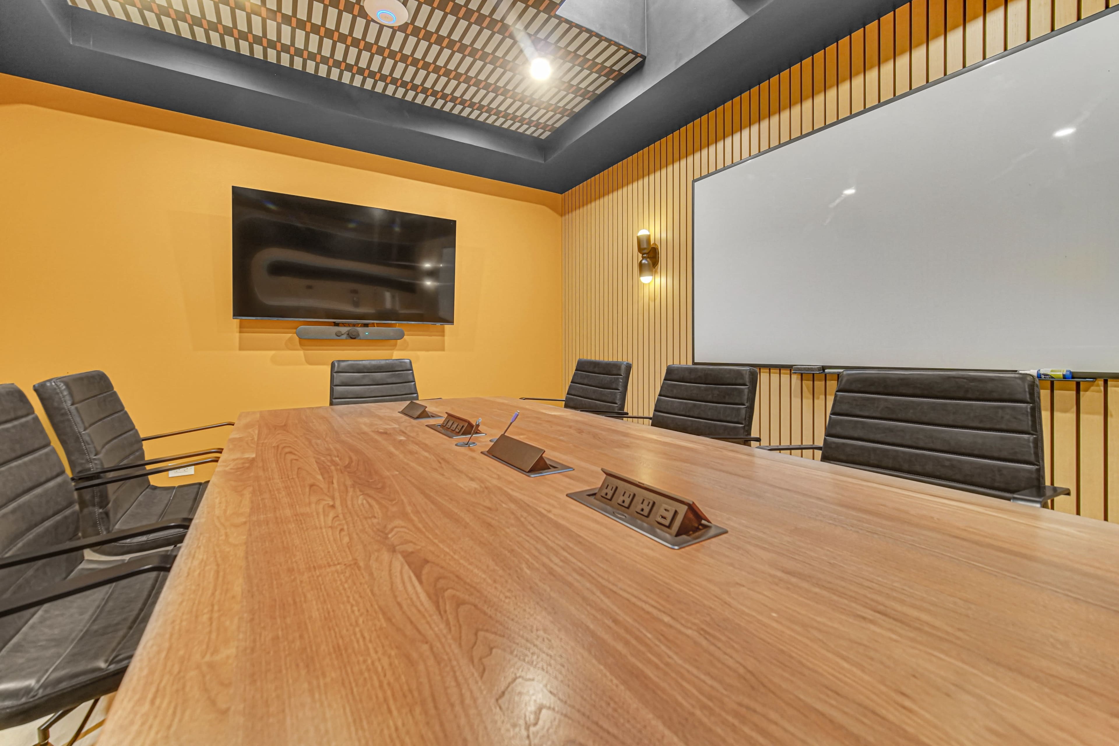The image shows a modern conference room with a large wooden table, black leather chairs, a mounted flat-screen TV, and a whiteboard on an orange wall.