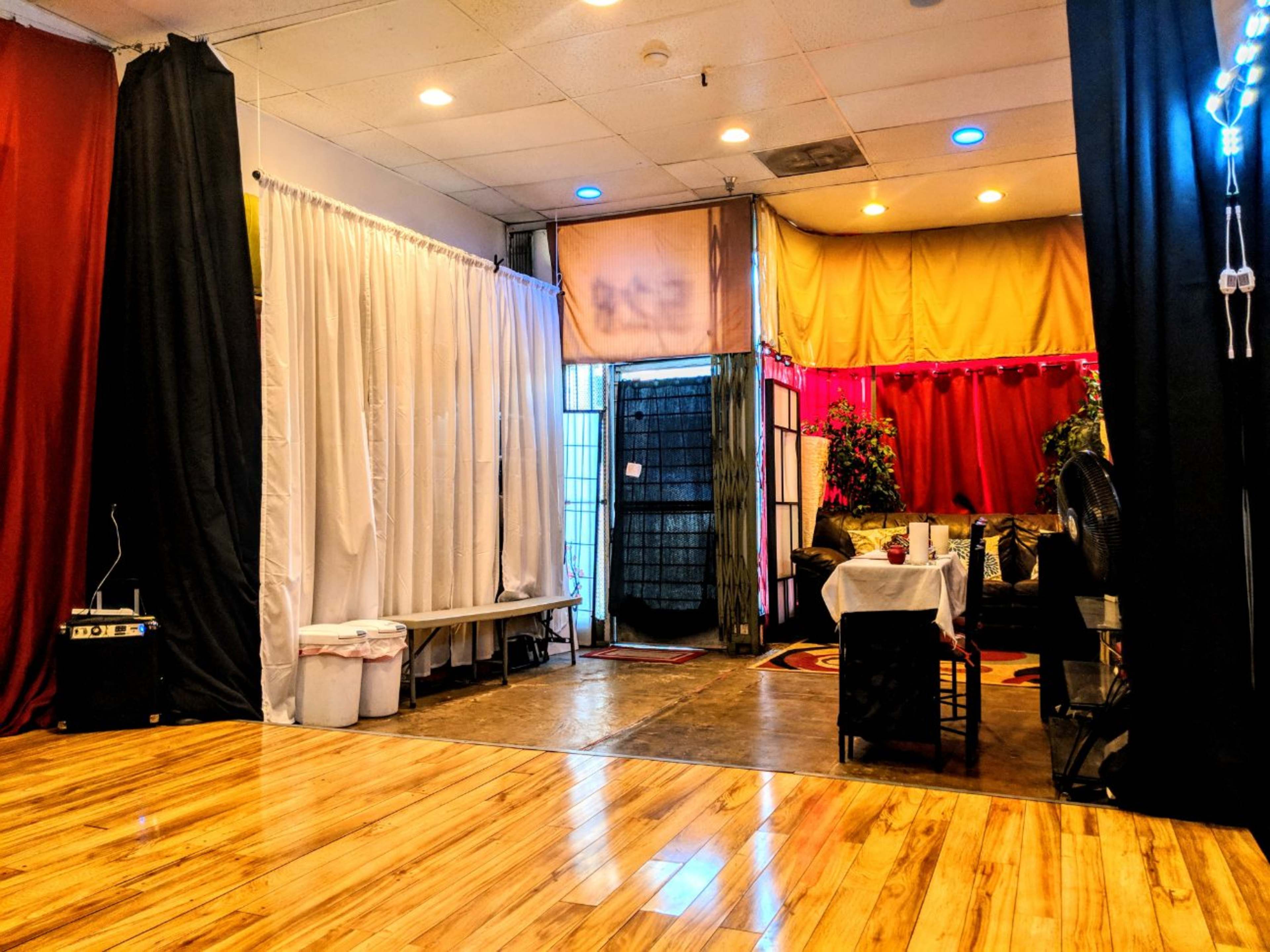 An indoor space divided by curtains, with wooden flooring, a table set up in the center, and various plants in the background.