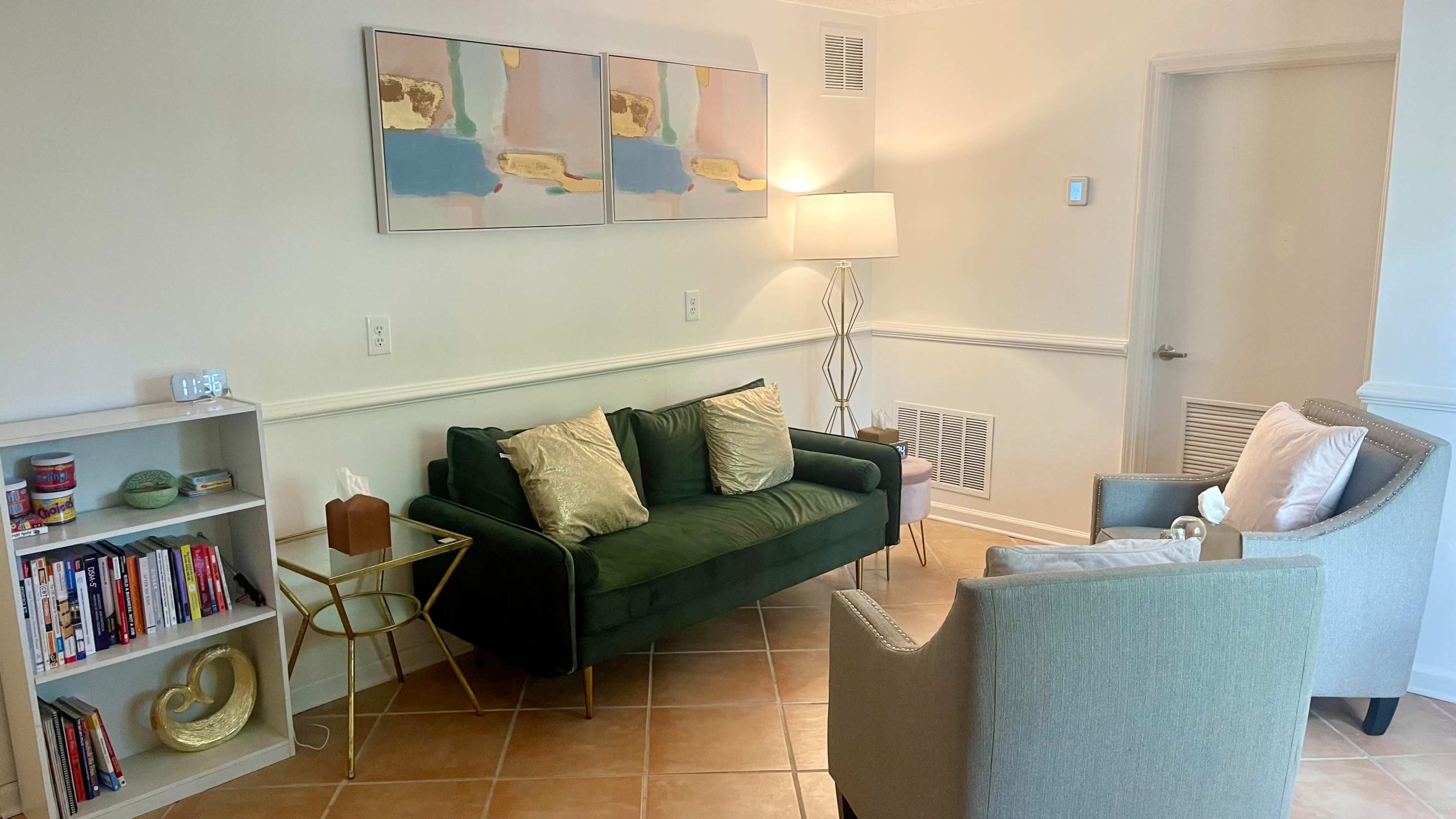 A living room features a green sofa with decorative pillows, a light-colored armchair, a small round table, a bookshelf filled with books, and abstract artwork on the wall.