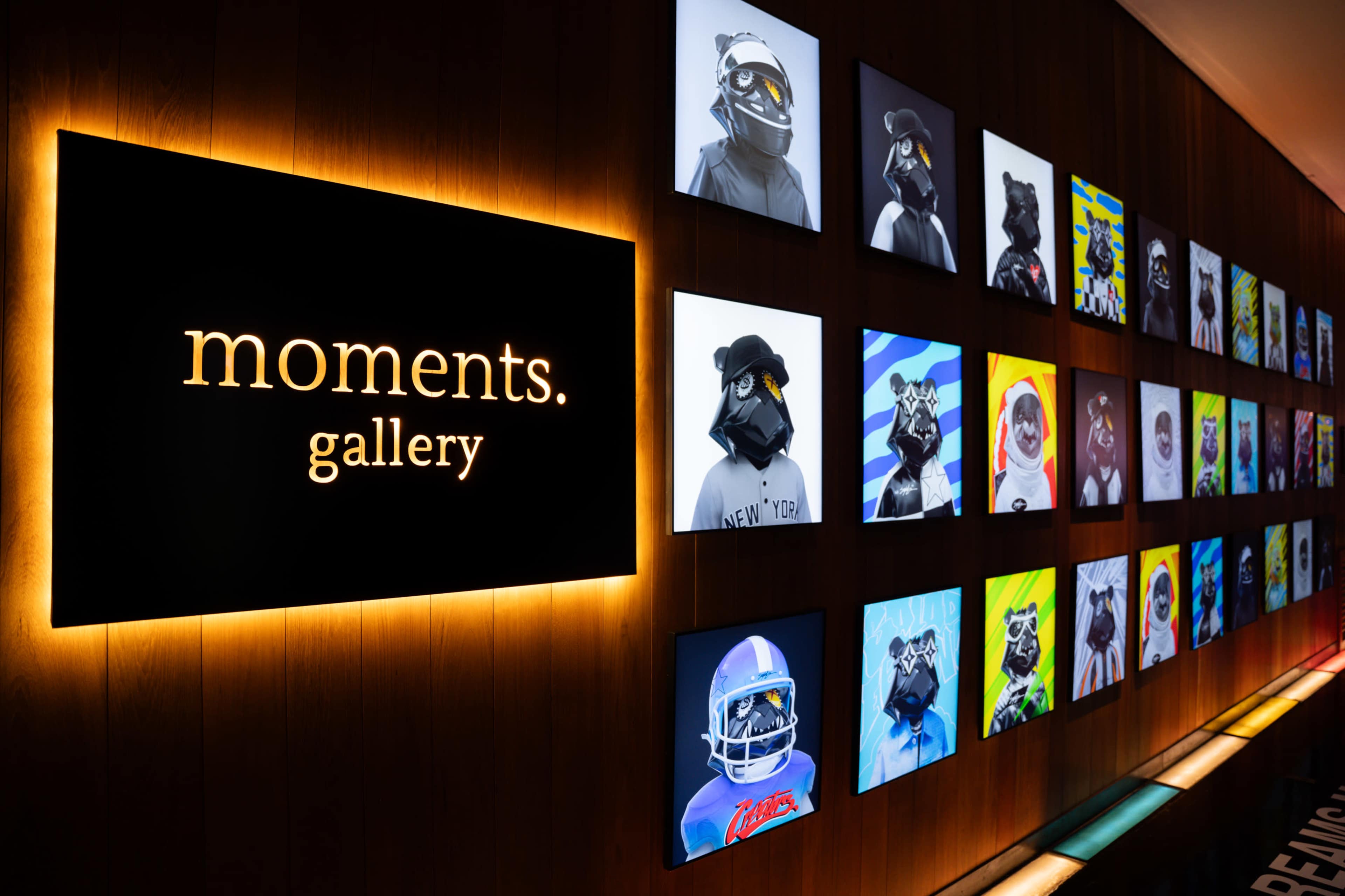 The image shows a gallery wall with a large illuminated sign reading "moments. gallery" and several square art pieces featuring stylized portraits of characters with masks and helmets.