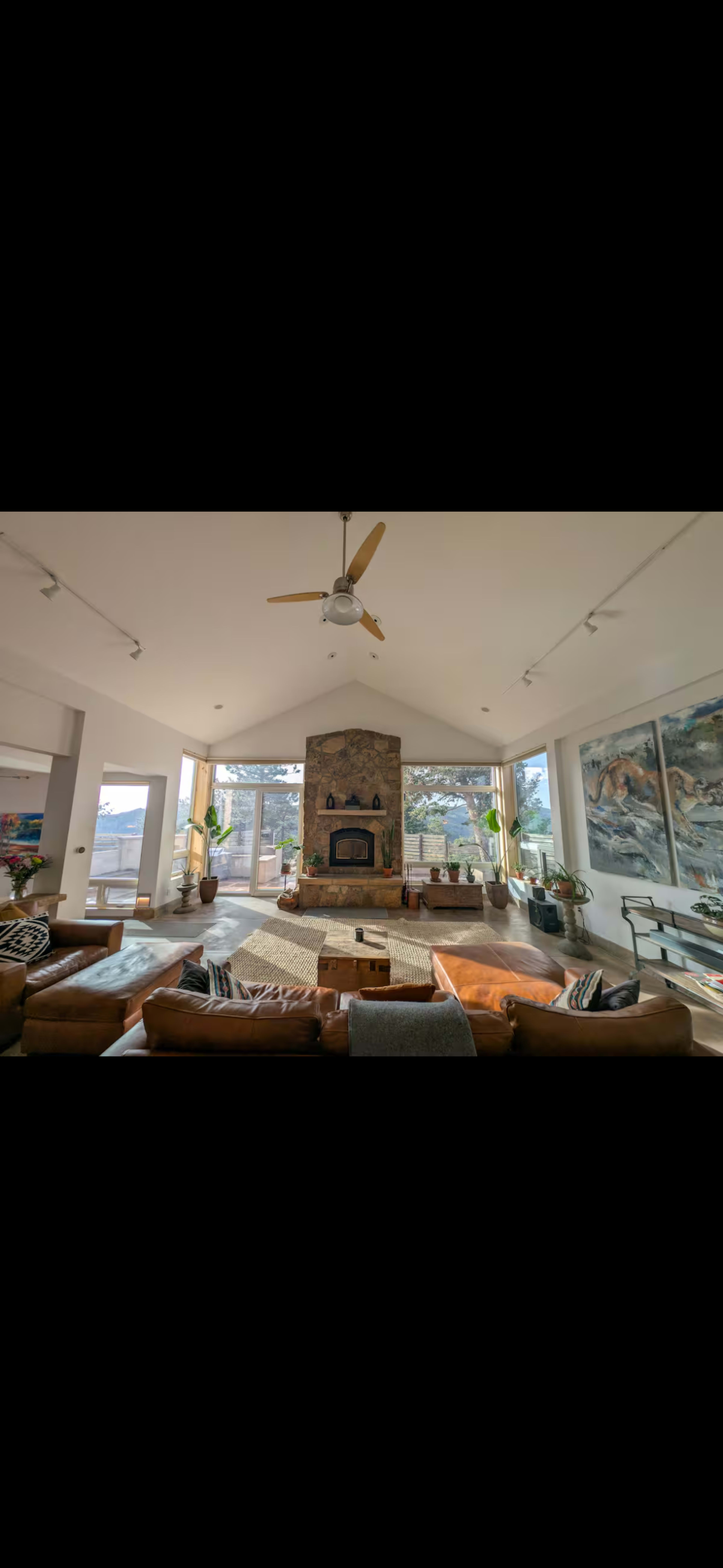 The image shows a spacious living room with a large stone fireplace, leather seating, and large windows that offer a view of the outdoors.