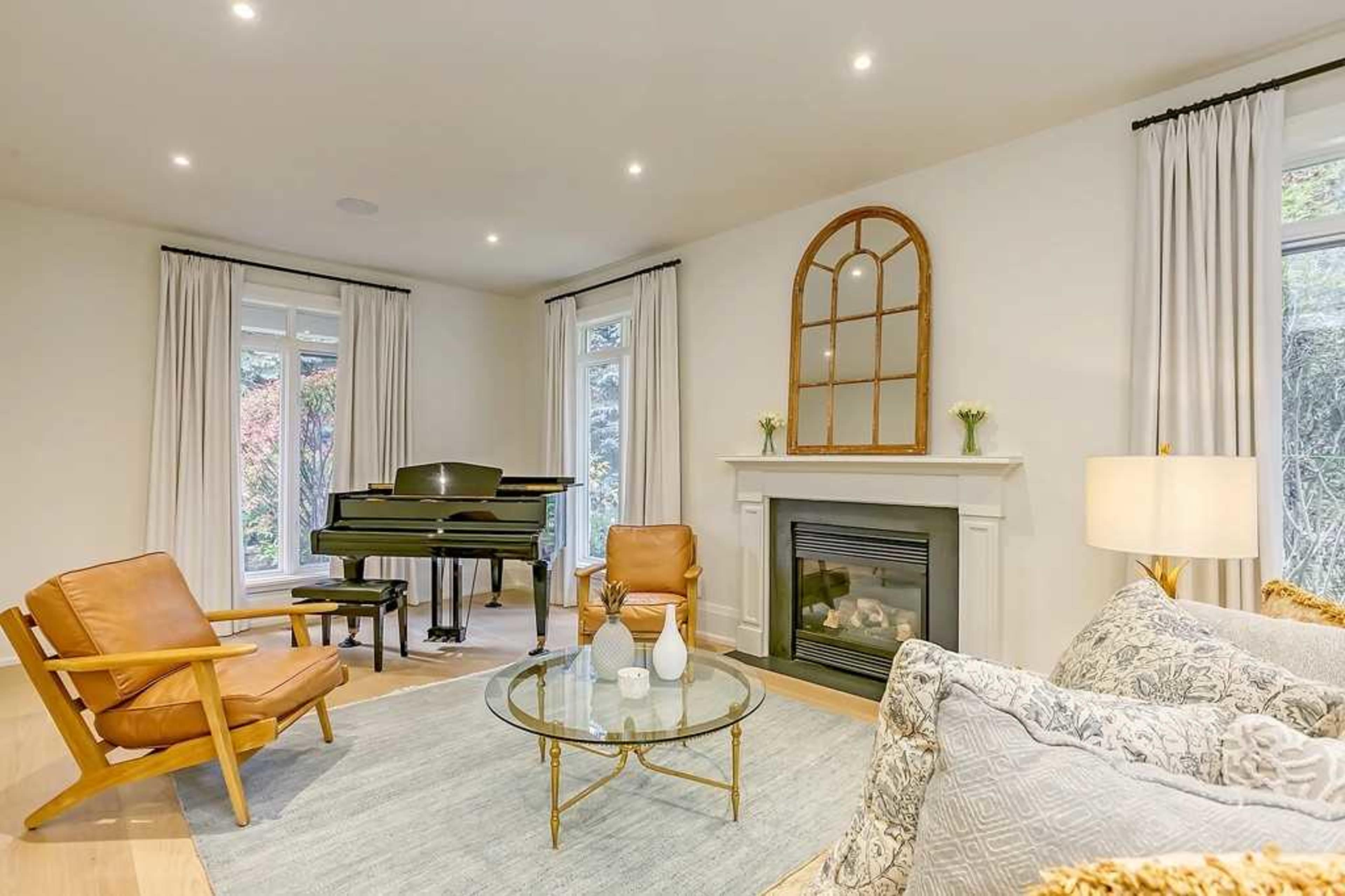 A stylish living room features a piano beside a fireplace, with two chairs, a glass coffee table, and large windows allowing natural light.