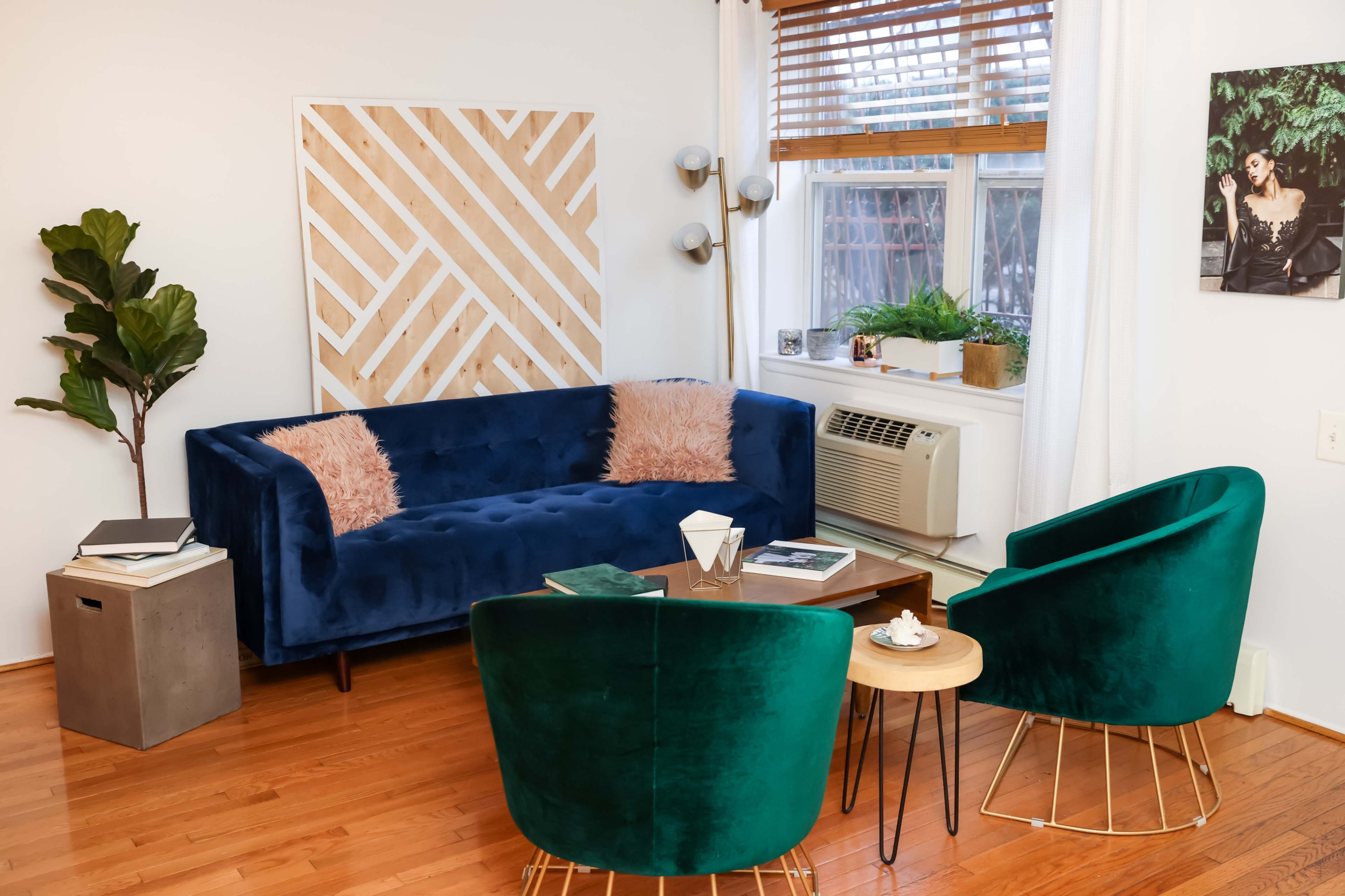 A blue velvet sofa and two green velvet chairs are arranged around a small table in a well-lit living room with wooden flooring and large windows.