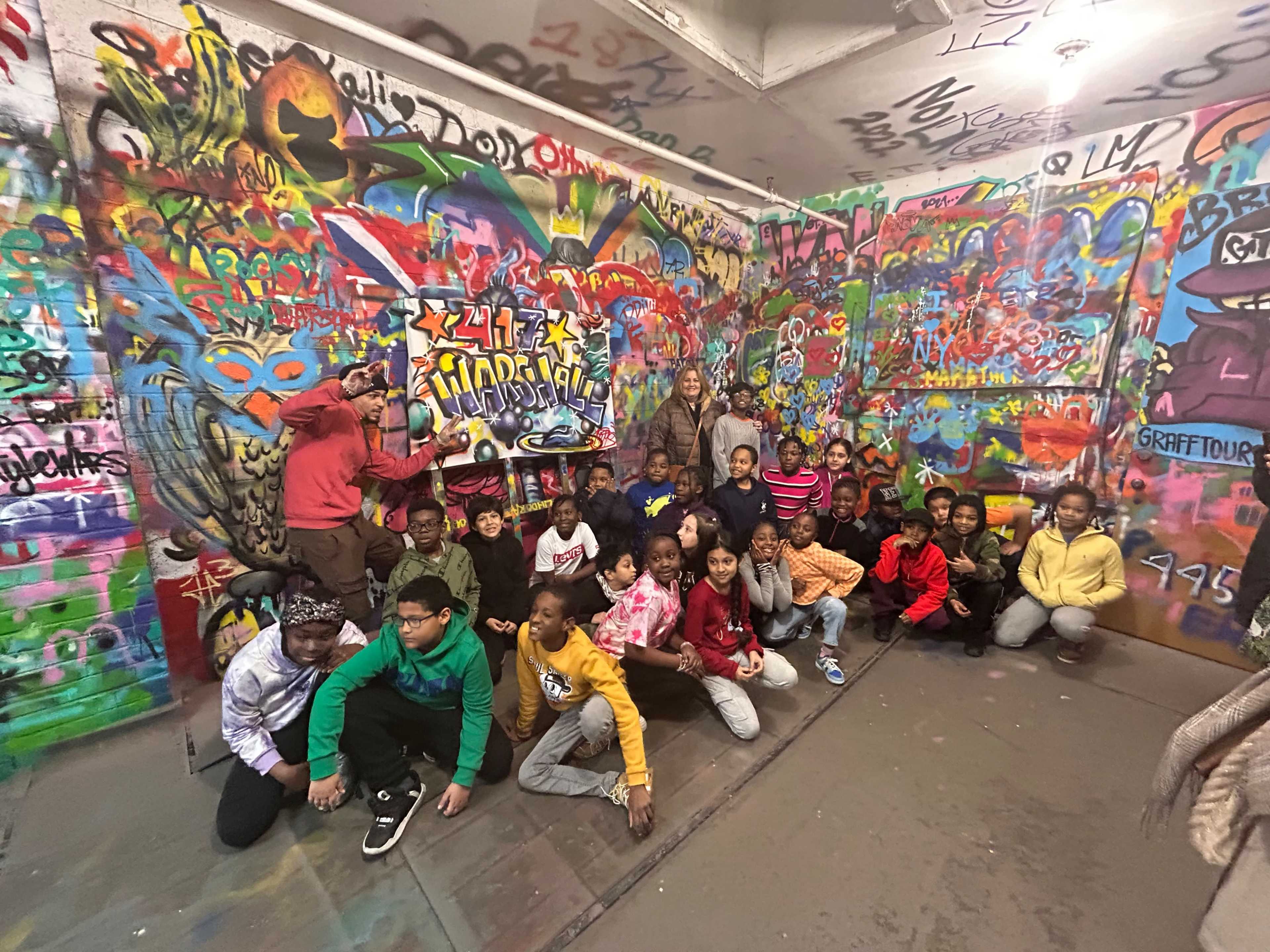 A group of children poses in front of a colorful, graffiti-covered wall in an indoor space.