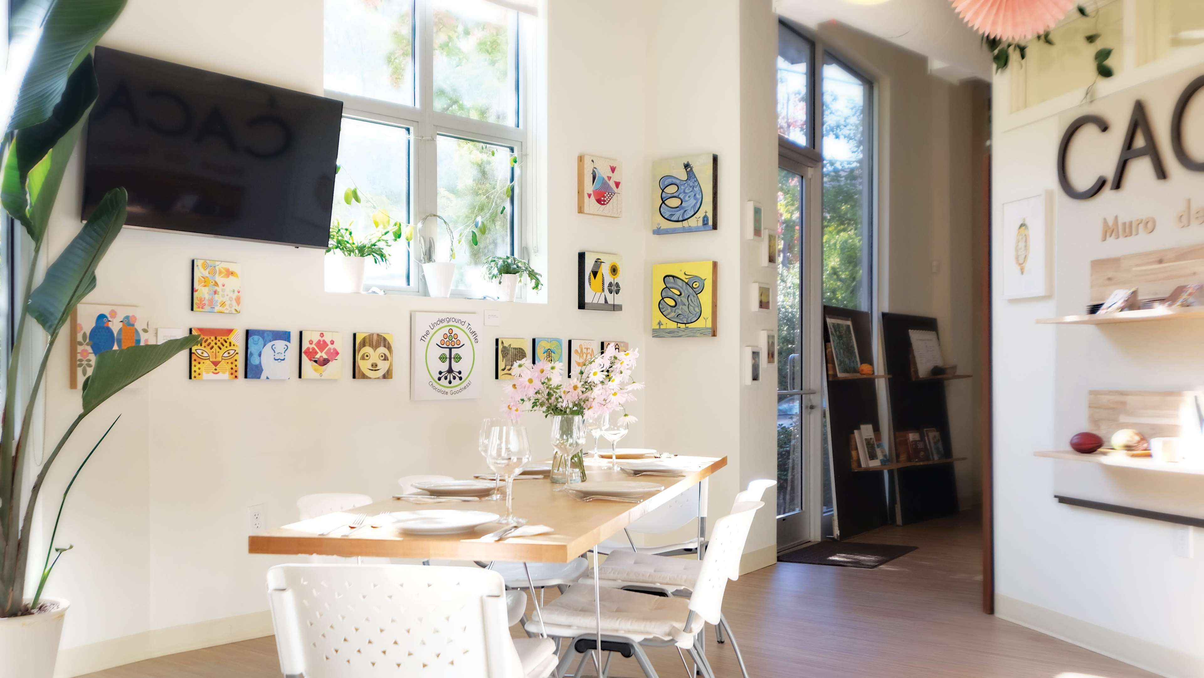 A brightly lit café interior with a wooden dining table set for two, decorated with flower arrangements, and walls adorned with colorful artwork.