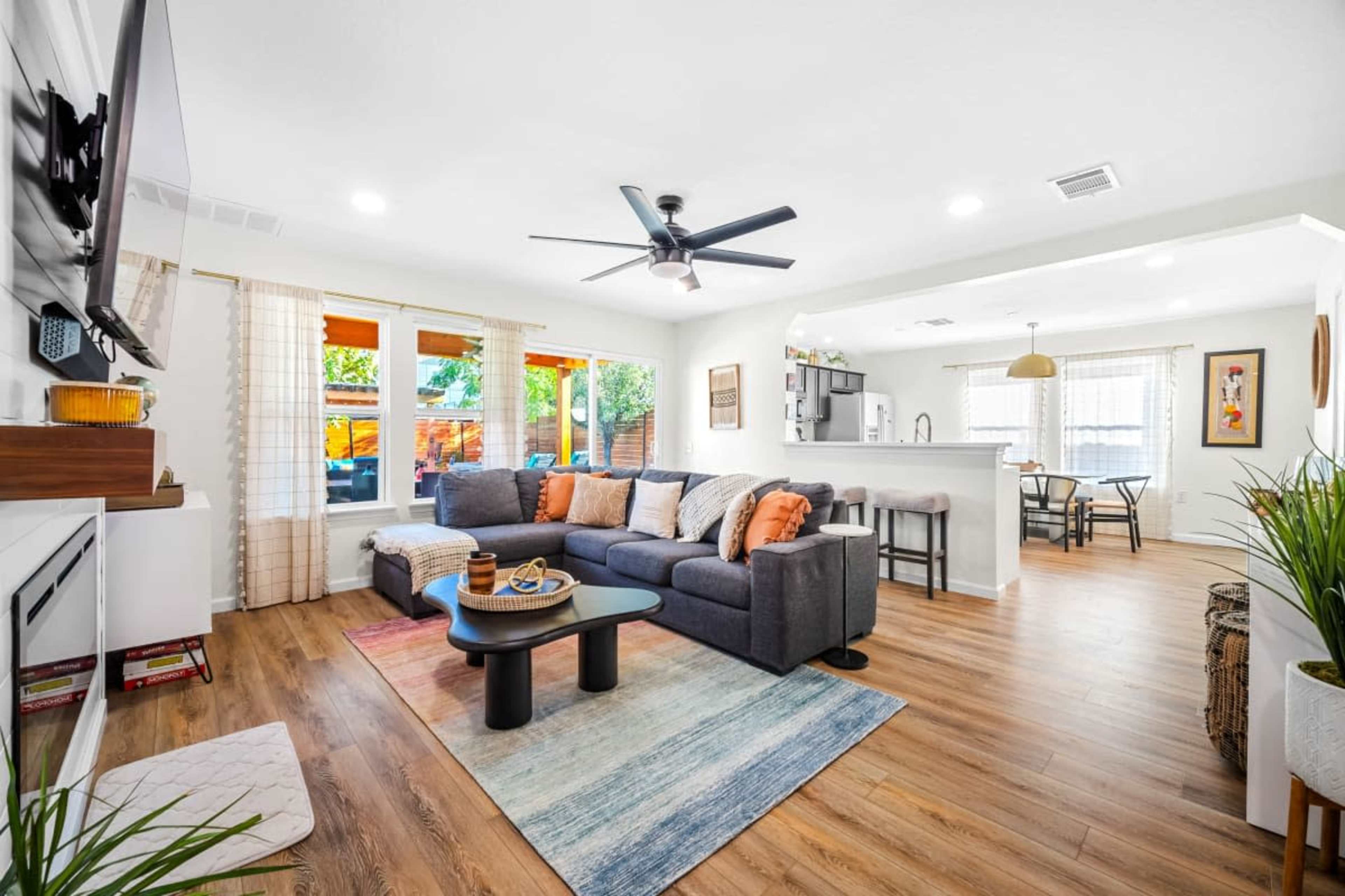 A modern living room features a large sectional sofa, a coffee table, and a television mounted on the wall, with a kitchen area and dining space visible in the background.