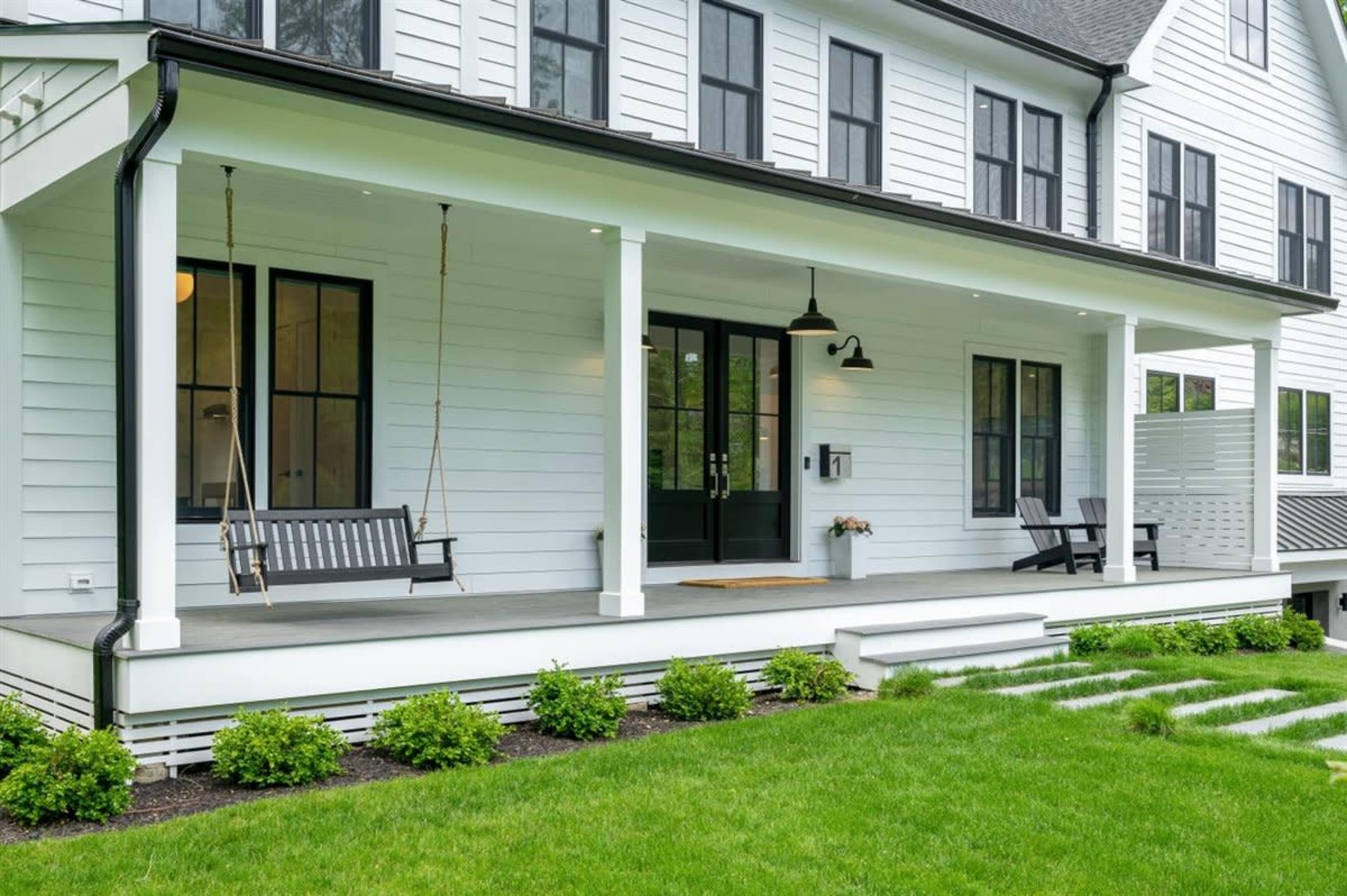 A modern house with a white exterior, featuring a large front porch with a swing, black accents, and a well-maintained lawn with neatly arranged stepping stones.
