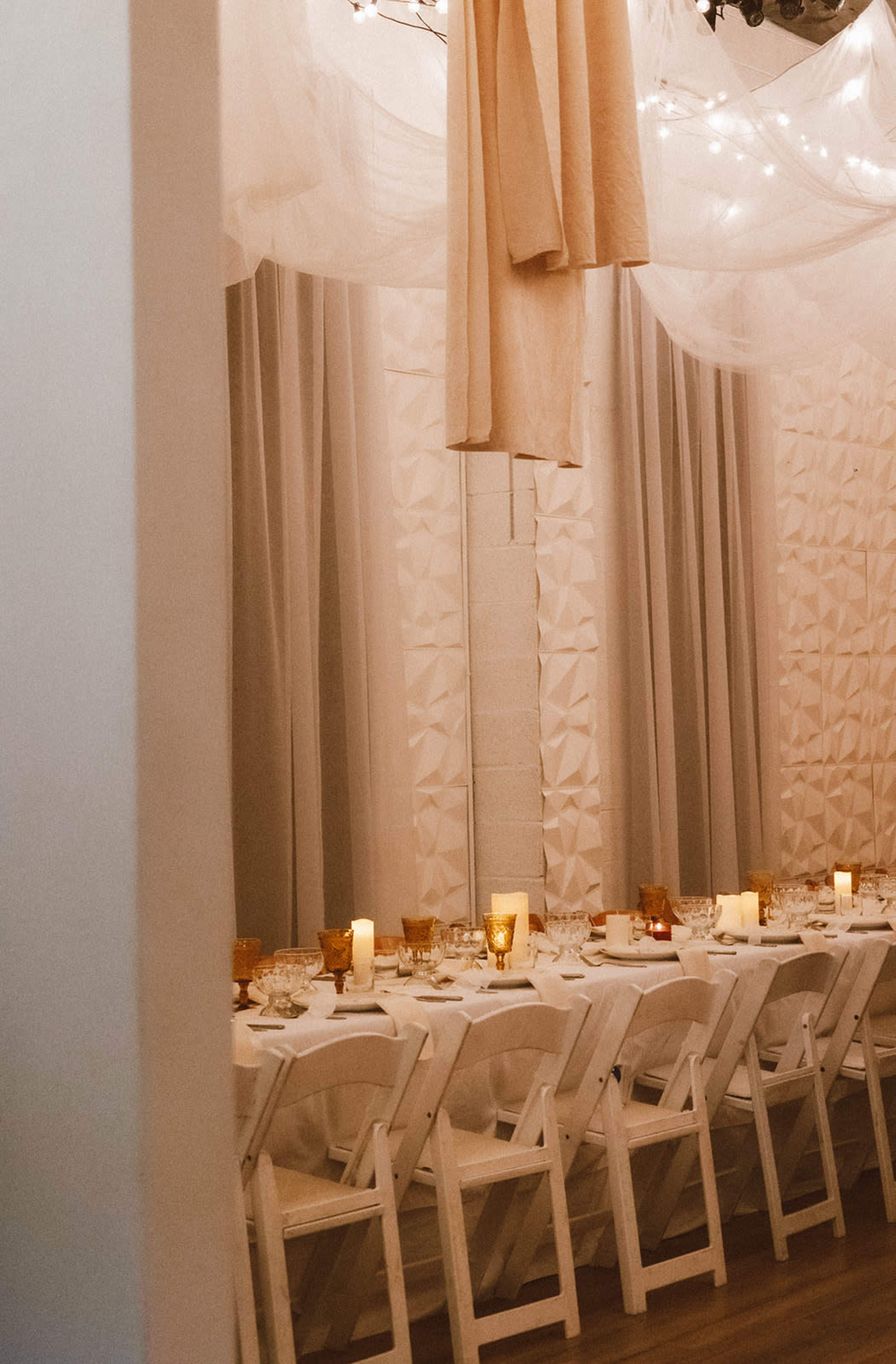 A decorated dining area features a long white table set with glassware and amber candles under soft draping and warm lighting.