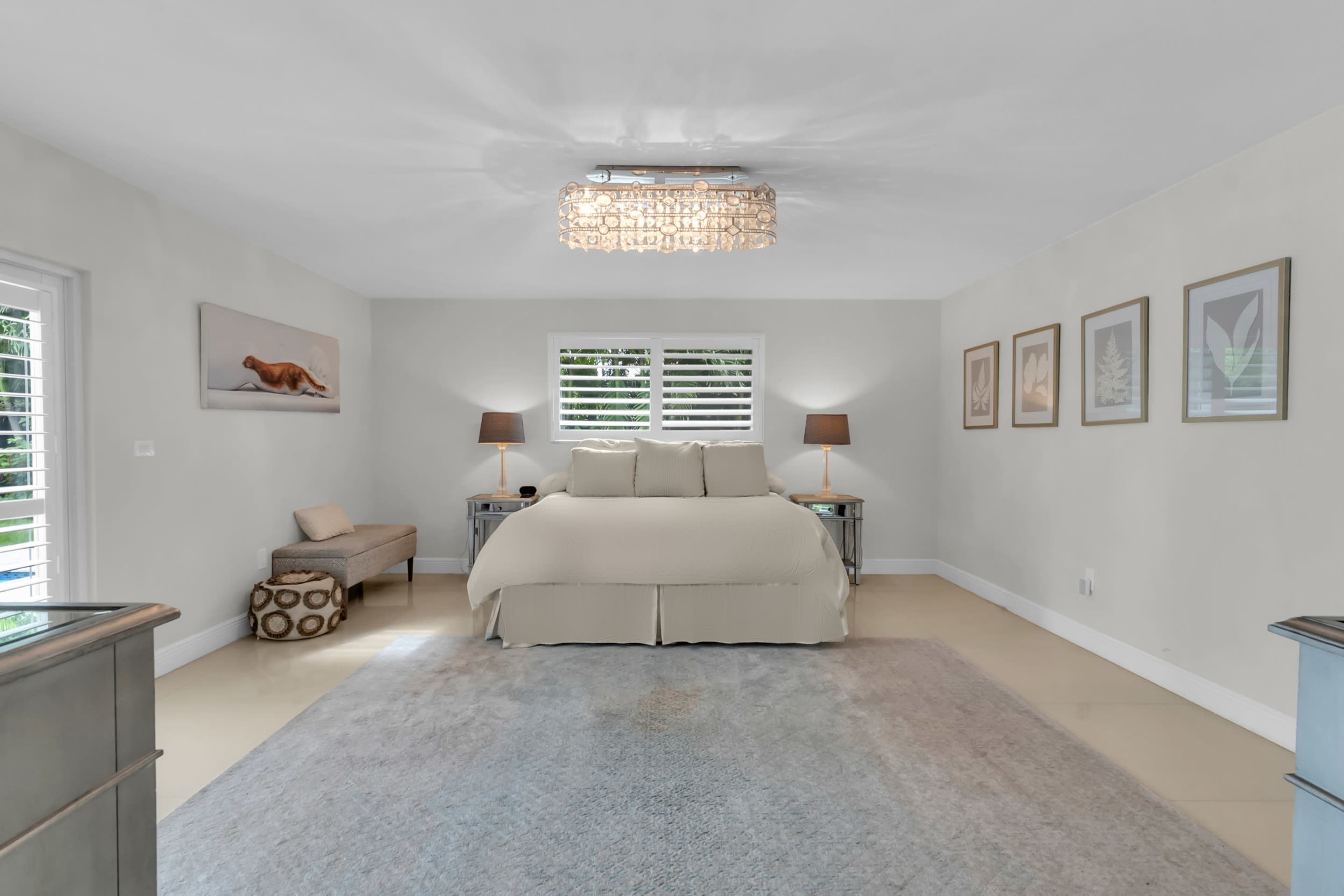 The image shows a spacious and well-lit bedroom featuring a large bed, two bedside lamps, a decorative bench, and framed artwork on the walls.
