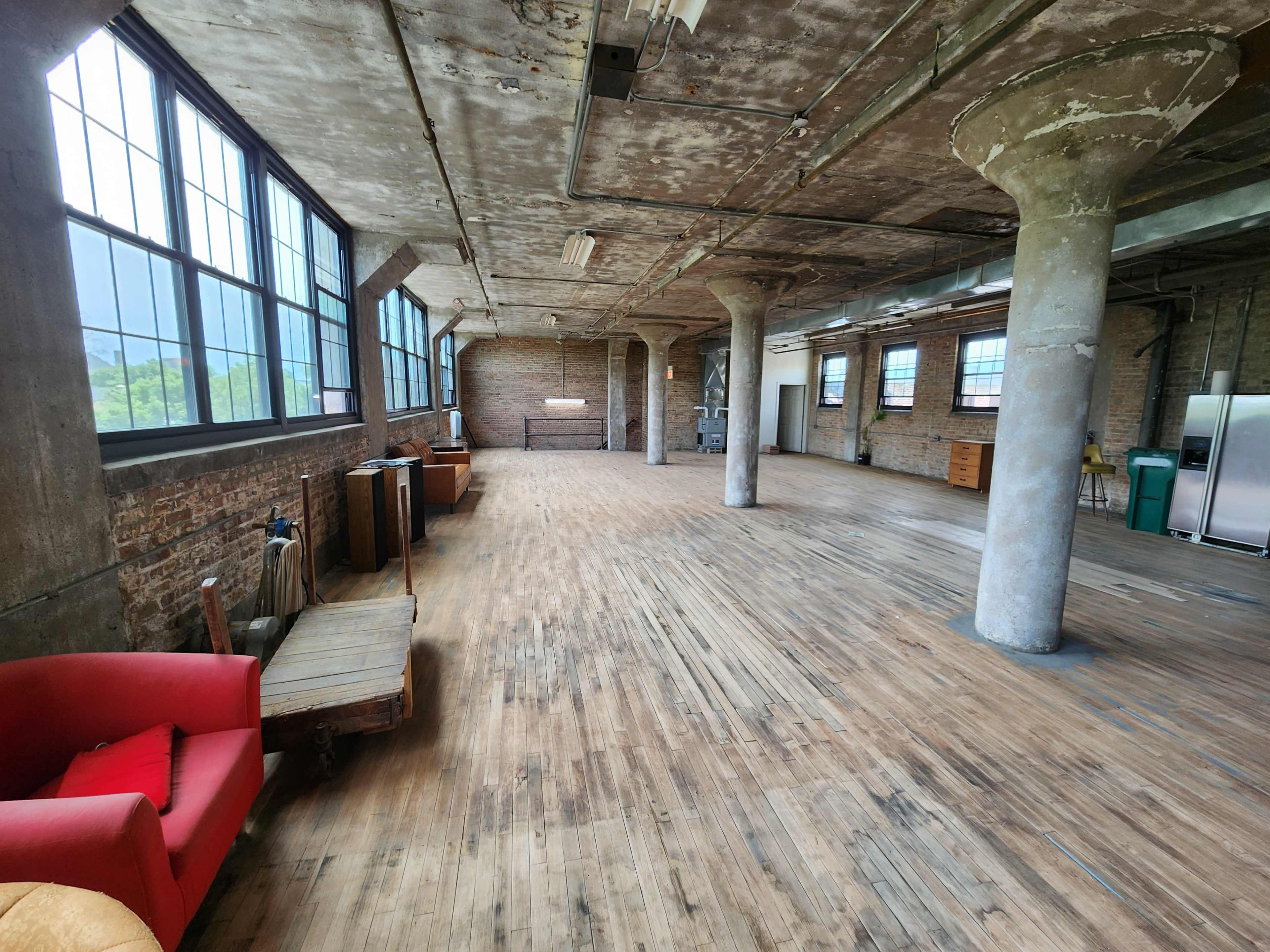 A spacious industrial-style room with large windows, exposed beams, and wooden flooring, featuring a red couch and various furniture against the walls.