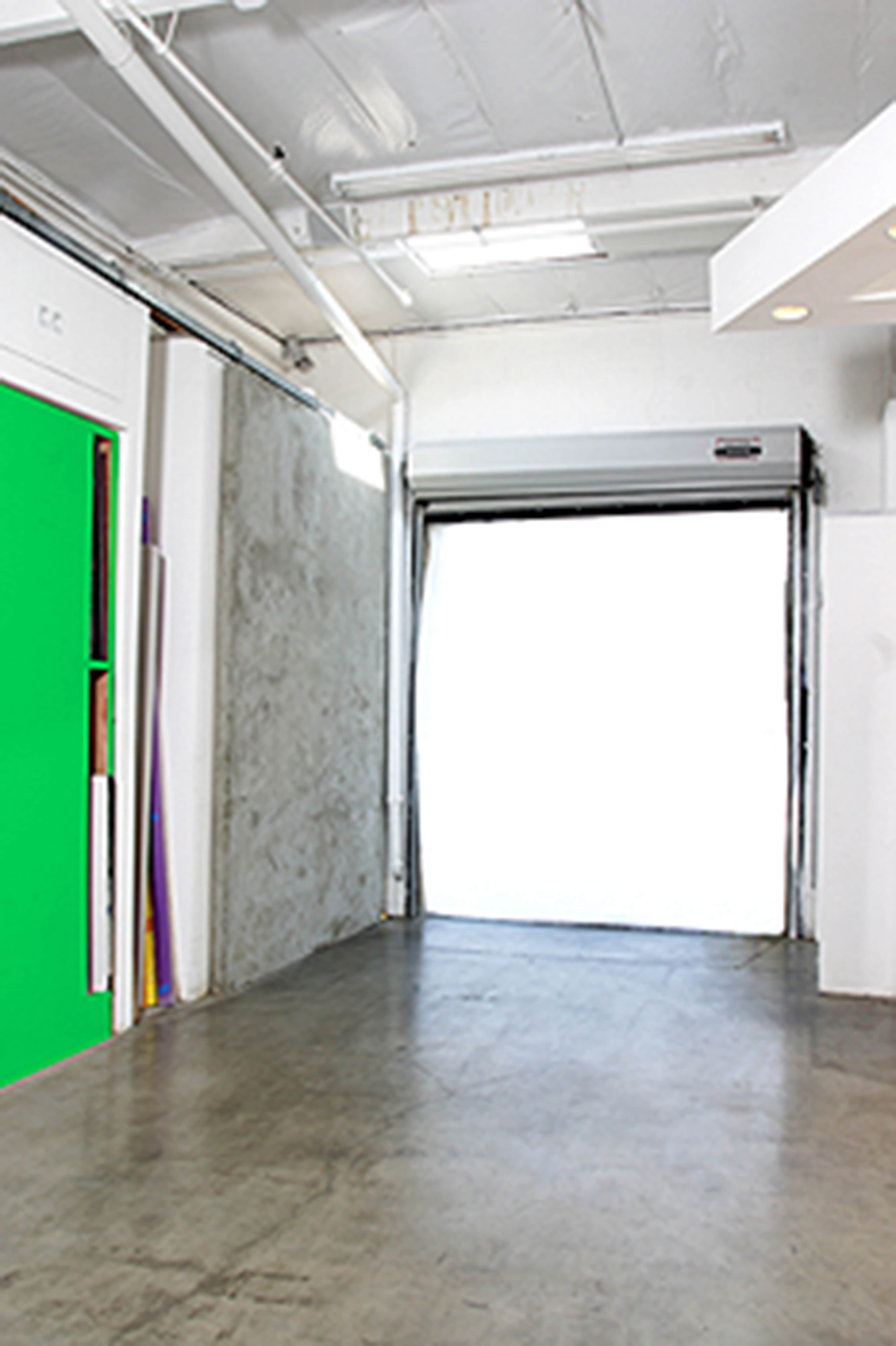 An empty industrial space with a green screen against one wall and a large open garage door at the far end.