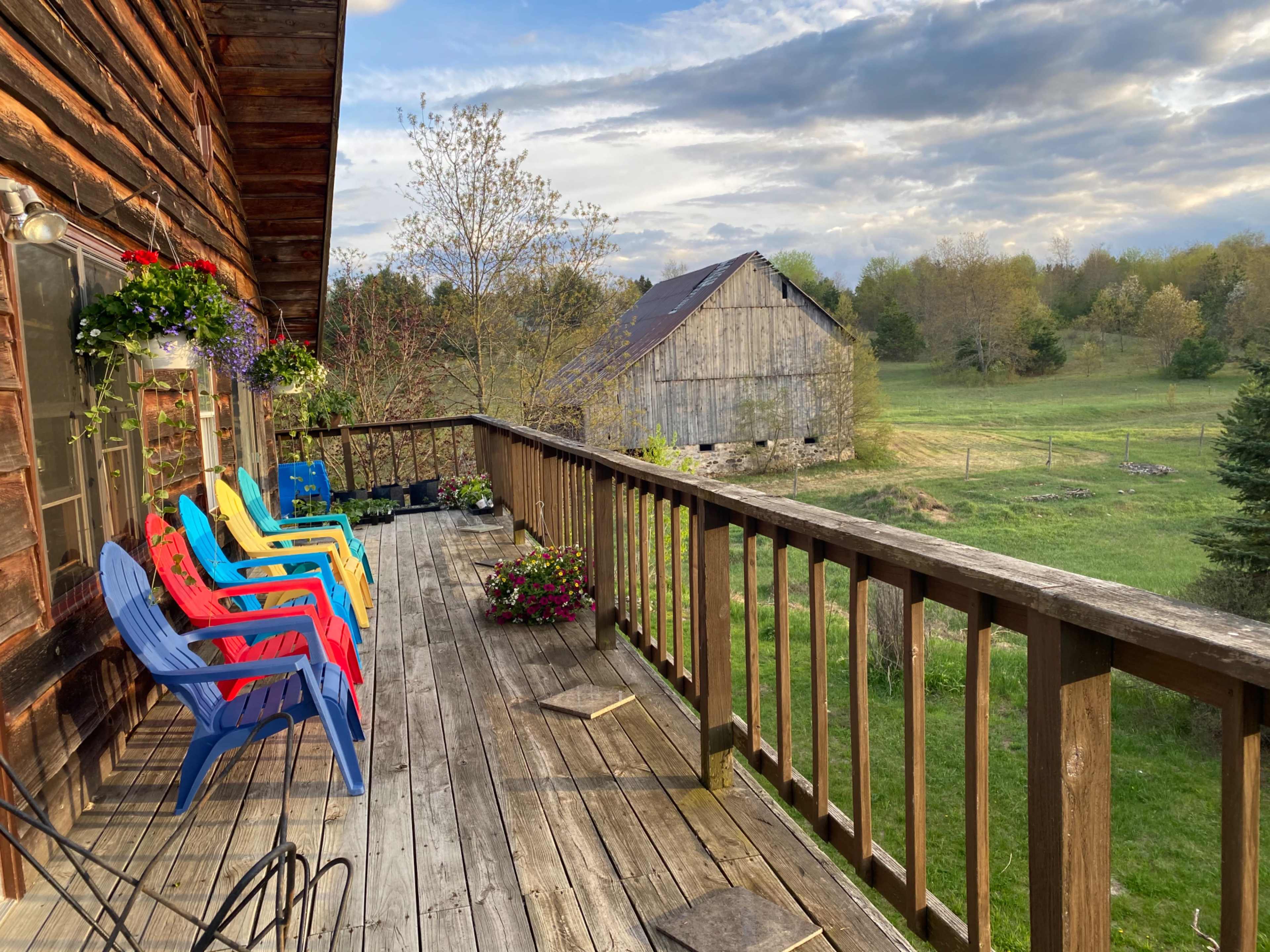 A wooden deck with colorful chairs overlooks a green field and a rustic barn in the distance.