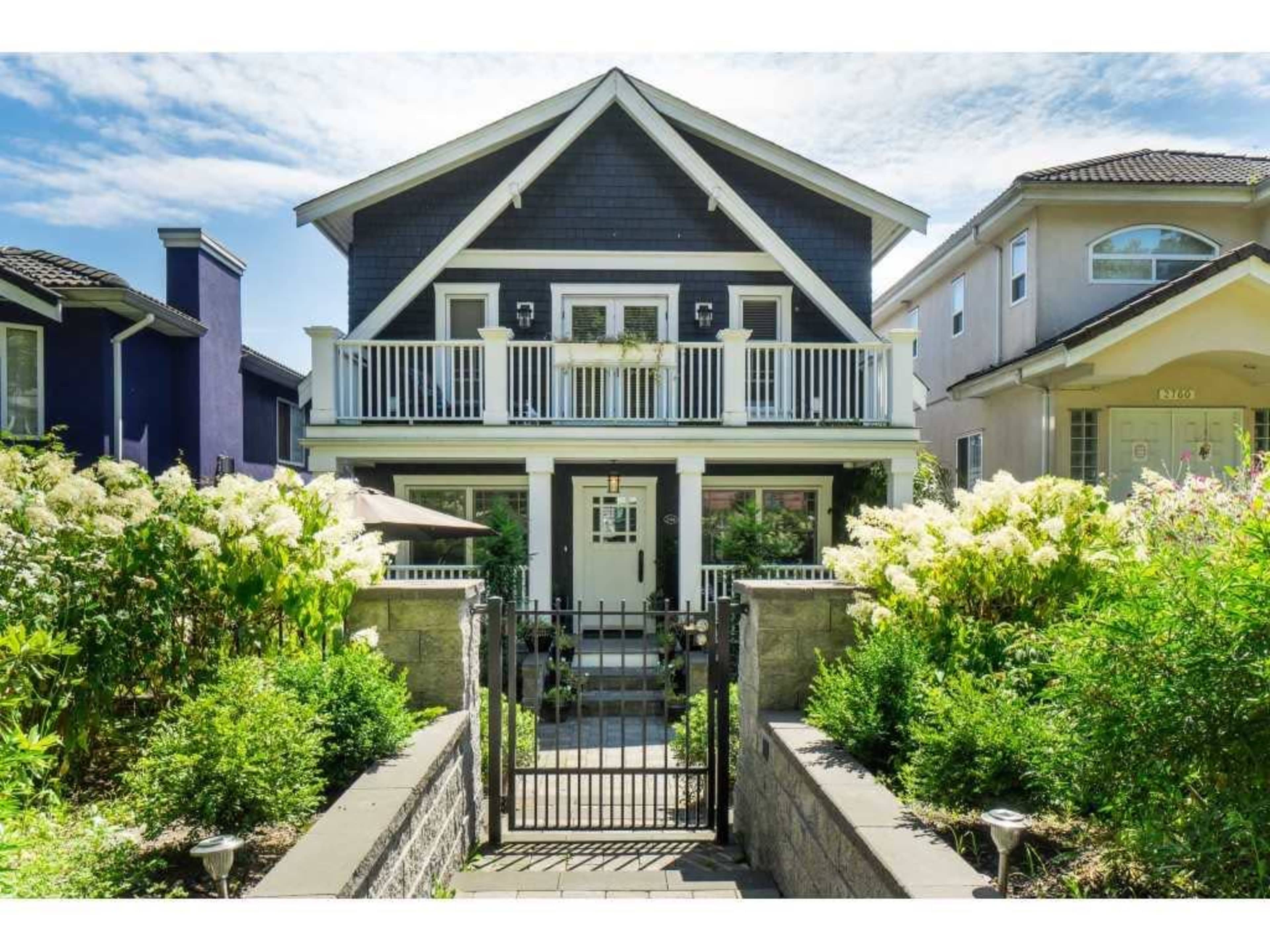 A two-story house with a dark exterior and white trim is framed by lush greenery and a front gate leading to a stone pathway.