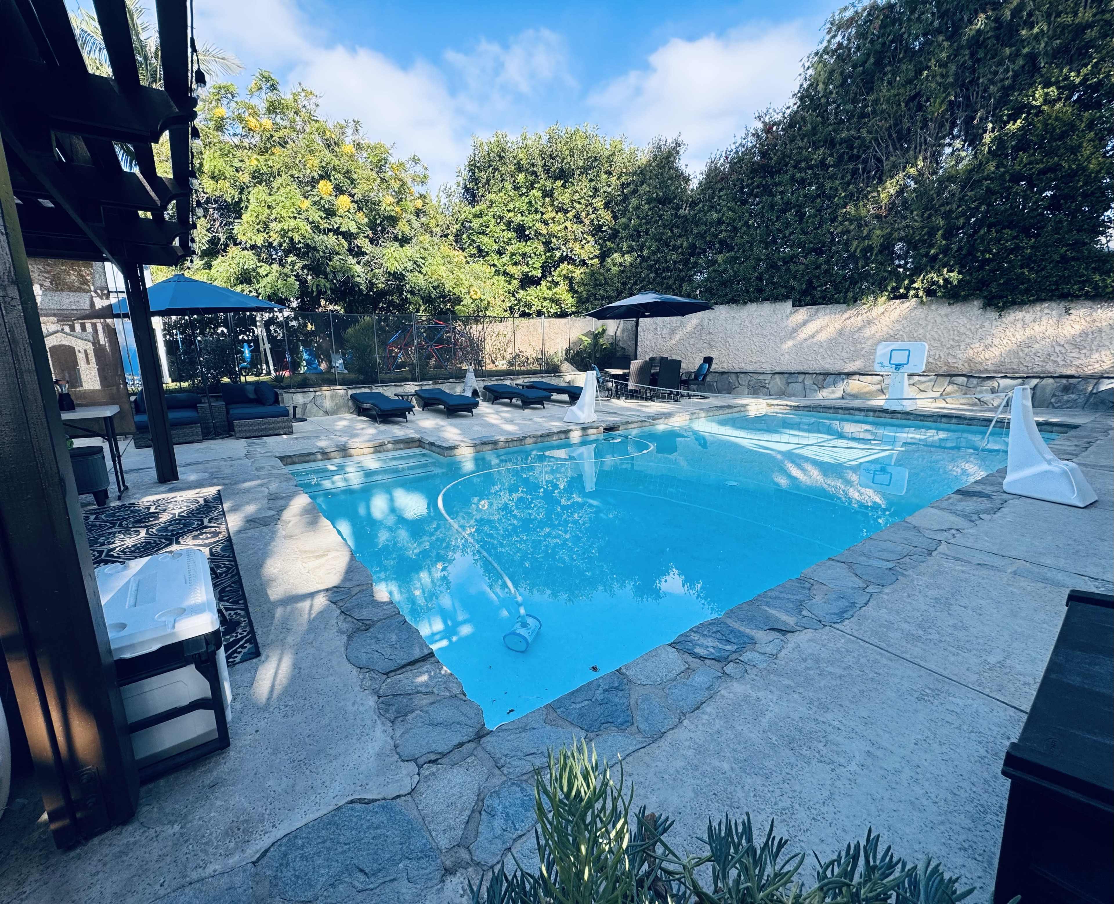The image shows a pool area with lounge chairs, umbrellas, and a surrounding stone patio in a residential backyard.