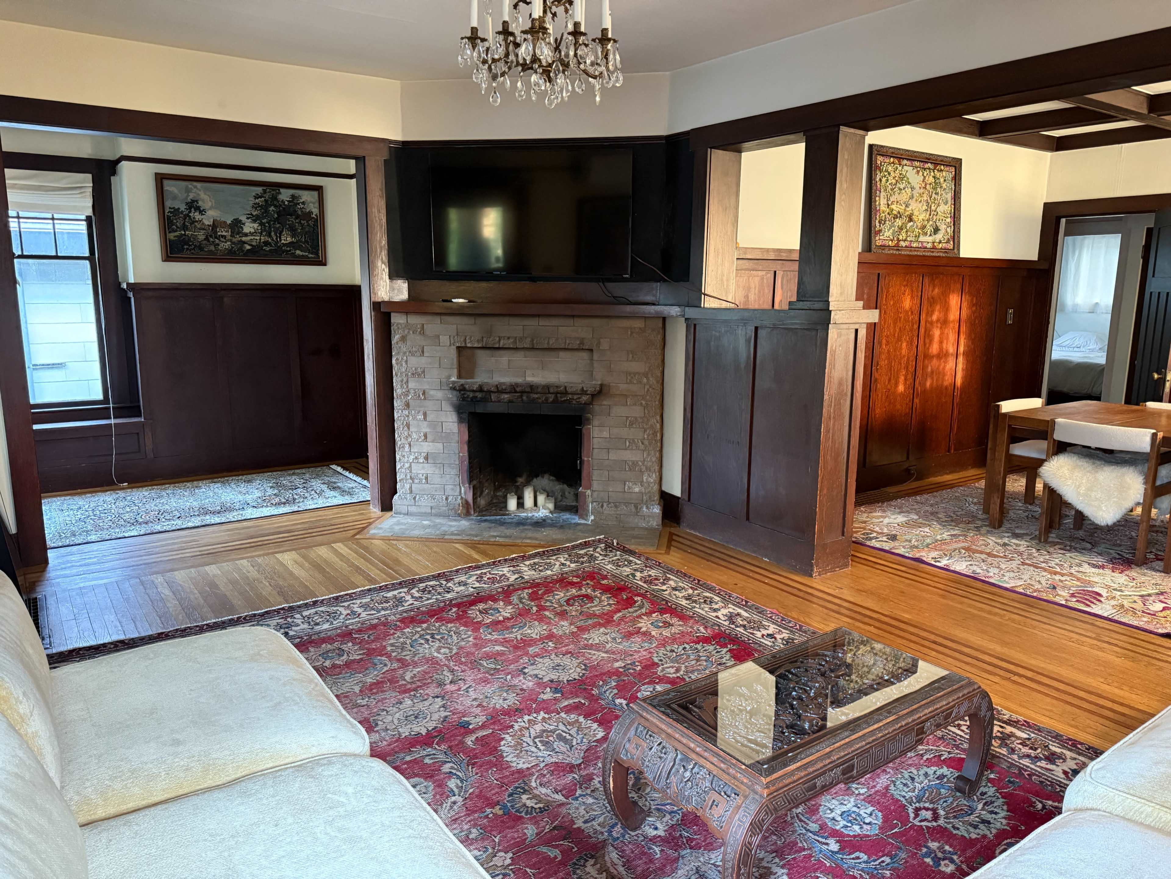 The image displays a living room with a fireplace, a large television mounted on the wall, a cream-colored sofa, and a wooden dining table surrounded by chairs.
