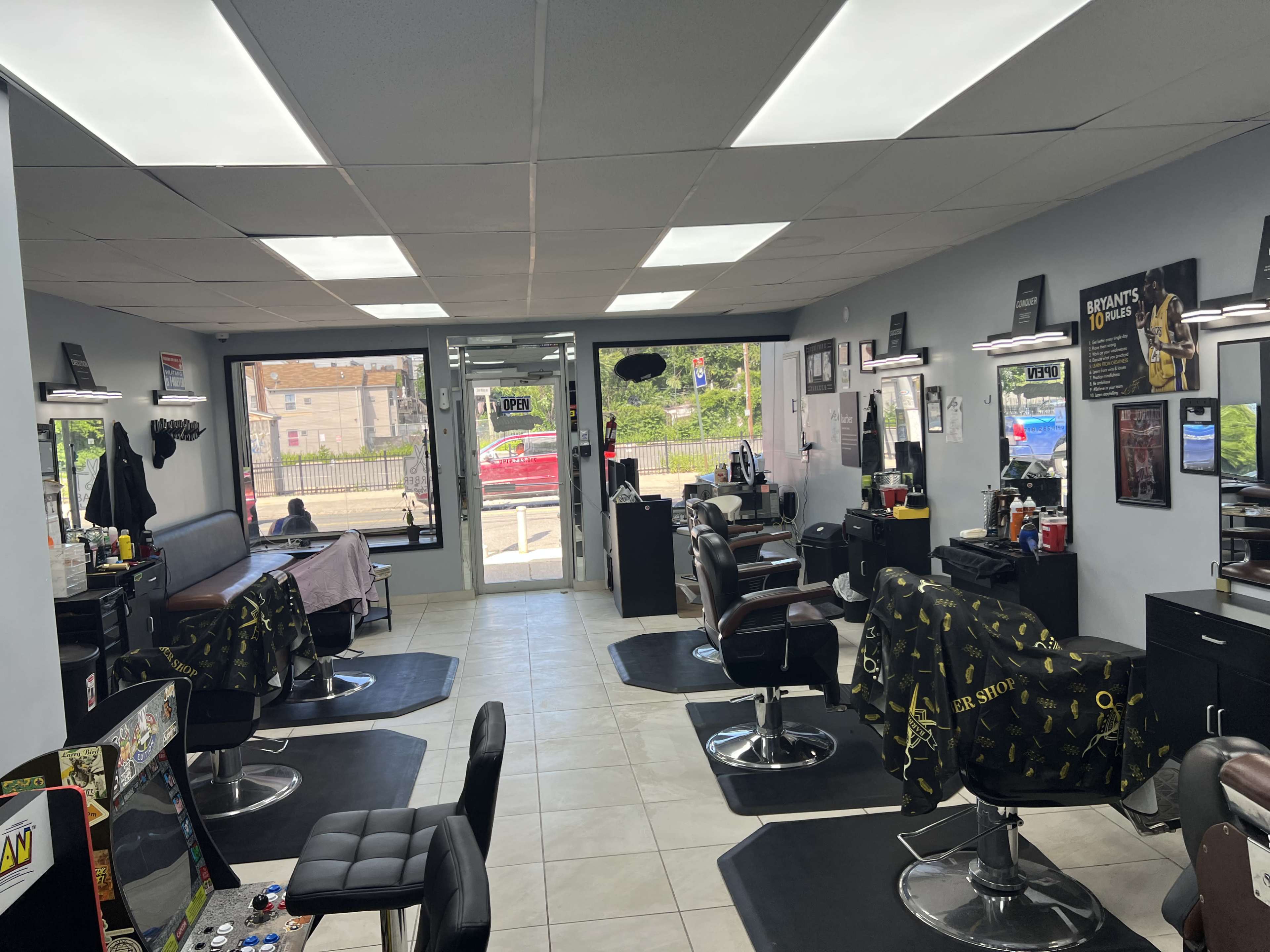 The image shows the interior of a barbershop with multiple chairs, mirrors, and equipment, along with large windows allowing natural light into the space.