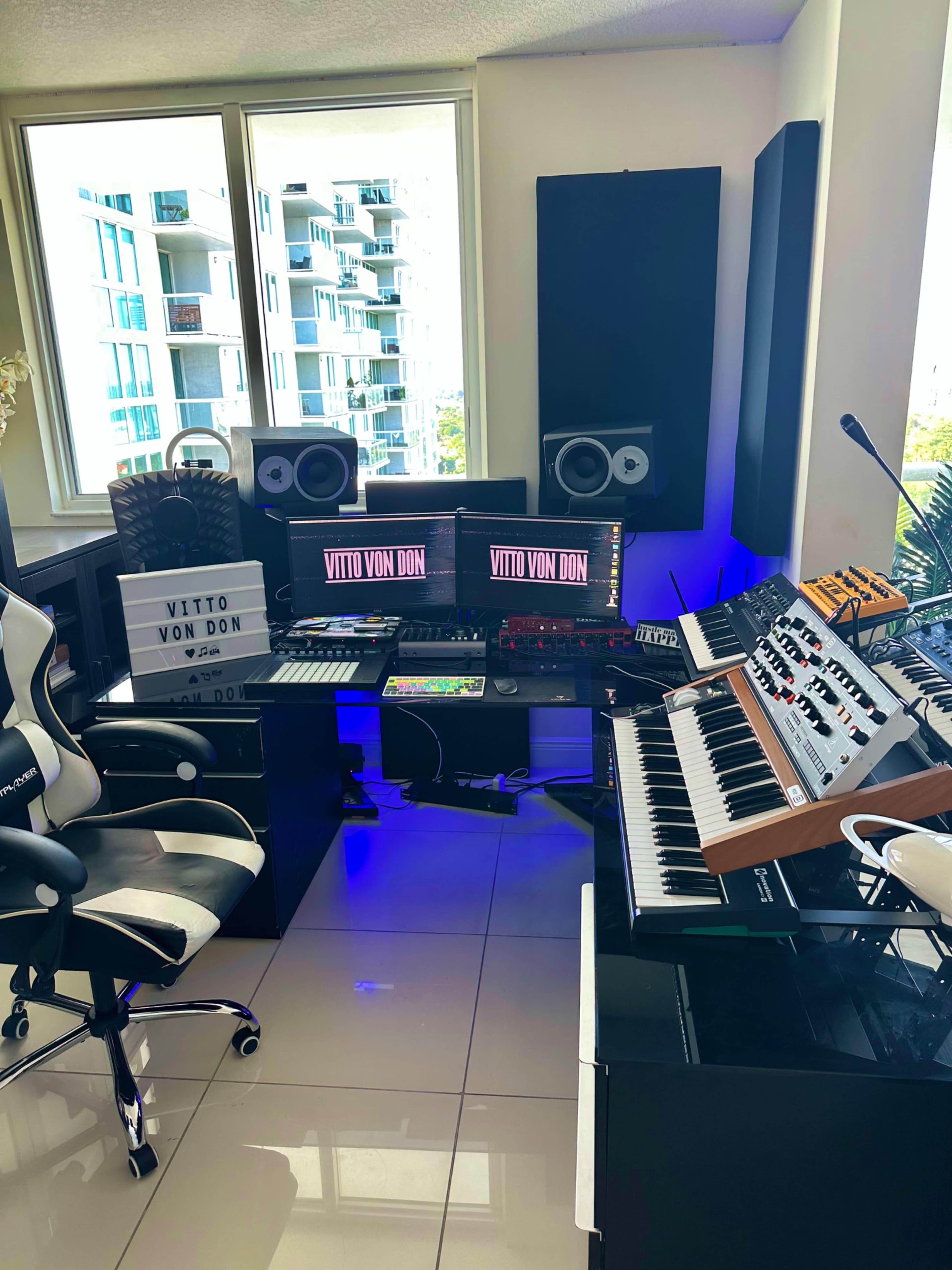 A modern music studio features multiple computer monitors, a tabletop synthesizer, a keyboard, and studio speakers, arranged around a swivel chair.