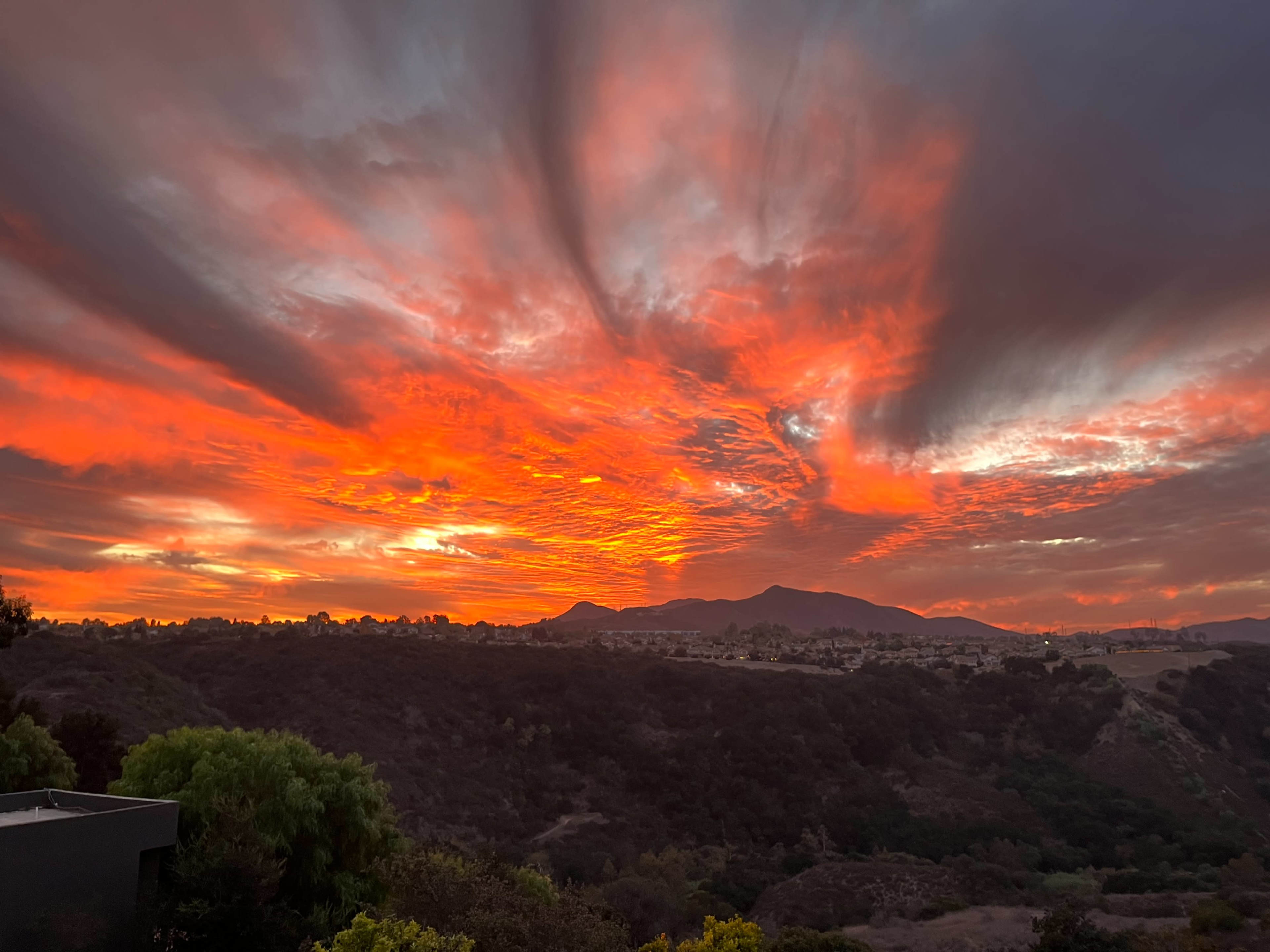 A vibrant sunset casts a fiery orange and red glow over a mountainous landscape, with dark clouds streaking across the sky.