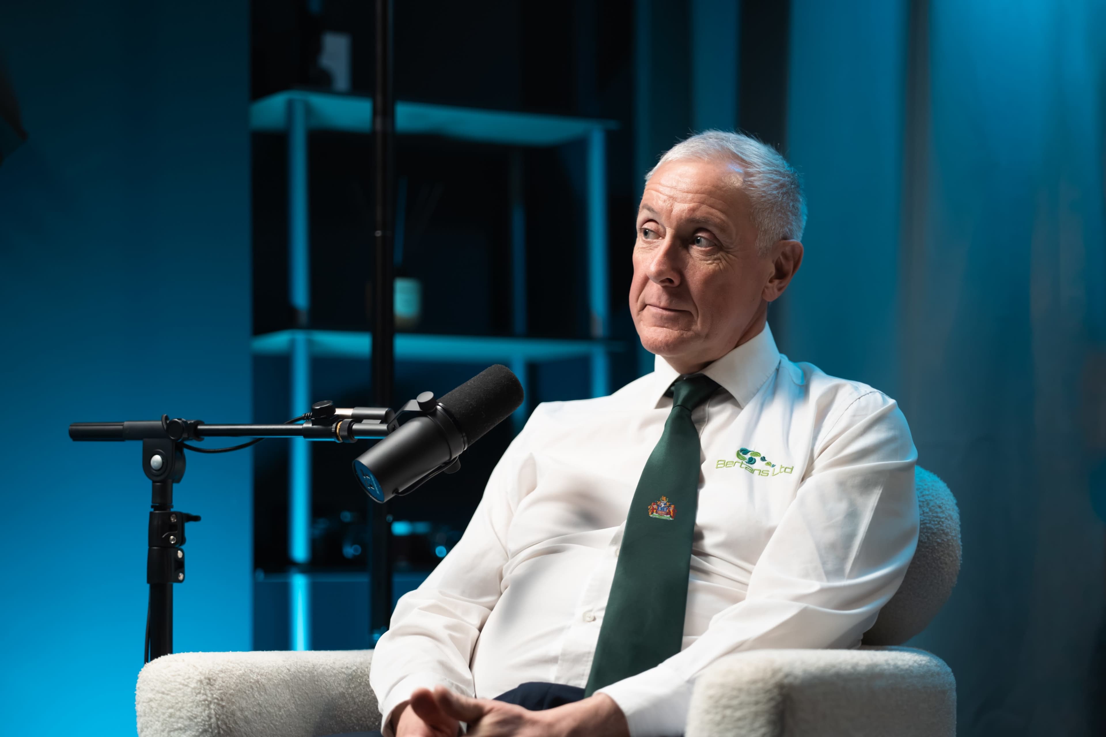 A man in a white dress shirt and green tie sits in a chair next to a microphone, with a blue-lit background and shelves in the distance.